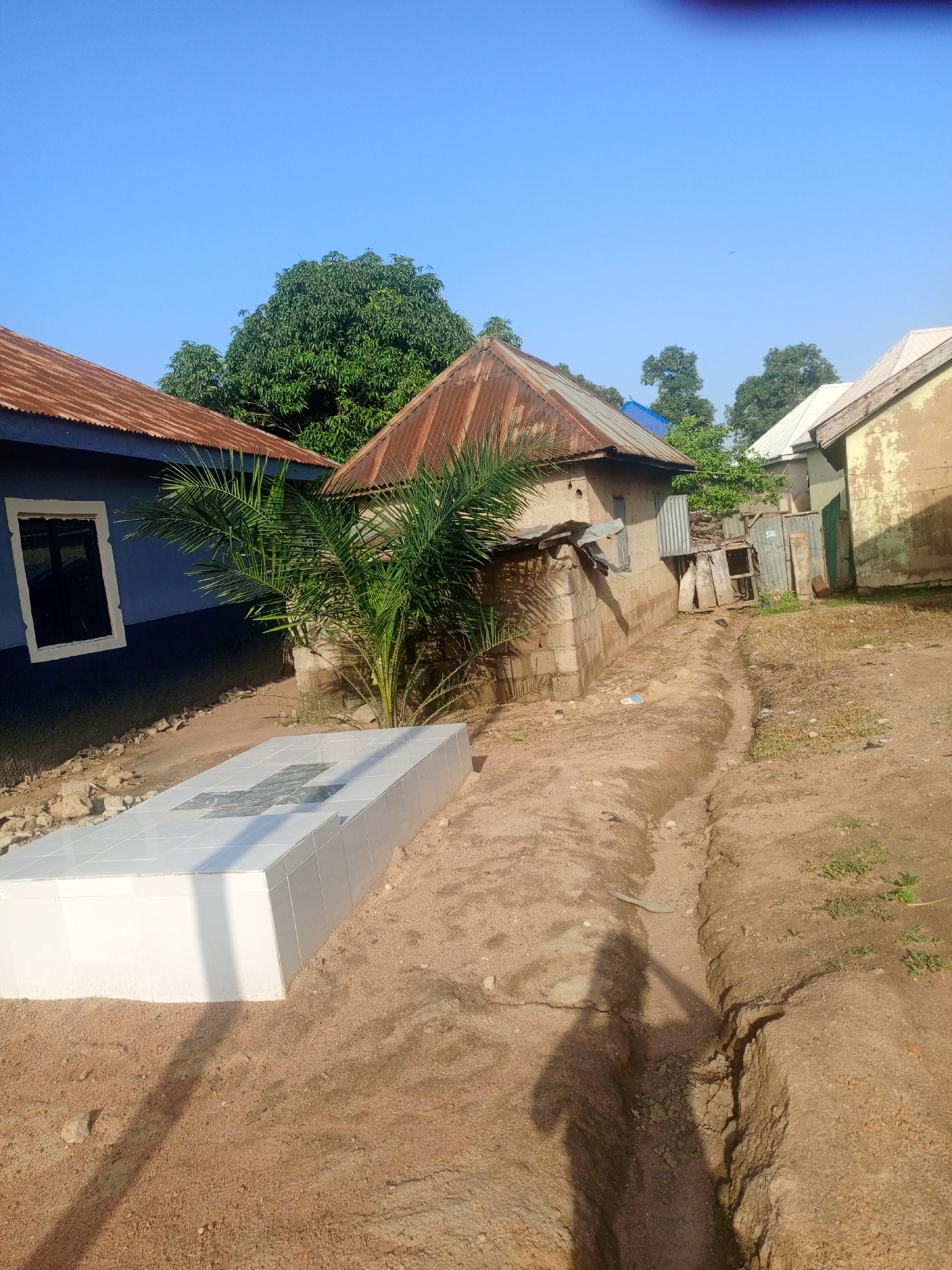 This image depicts an outdoor scene, likely in Jalingo, Nigeria, under a clear, bright blue sky, suggesting it is daytime with good weather. The foreground features a newly constructed white tiled tomb or grave, with a cross pattern visible on its surface. To the left, a blue-painted building with a rusted corrugated metal roof and a white-framed window is visible. Adjacent to the tomb, a small palm tree stands, and behind it are several small, rudimentary structures with corrugated metal roofs and walls constructed from concrete blocks and other materials, indicating a more rural or developing area. A dirt path or trench runs through the center of the image, with dried grass and some scattered debris. The shadow of a person holding what appears to be a tool, possibly a hoe or shovel, is cast across the path, suggesting someone is working or present in the area. The overall impression is one of a serene, possibly somber, but also lived-in environment, with a blend of new construction and older, more weathered structures. There is no visible text in the image.