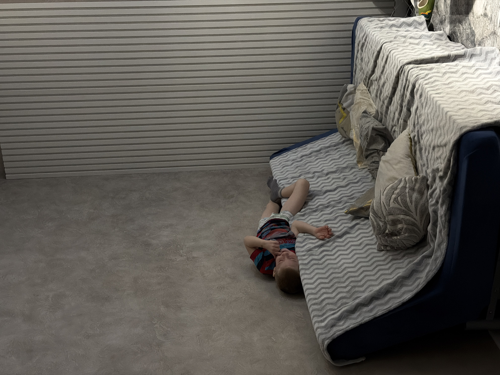 A child is lying on the floor next to a blue sofa in Rossiyskiy, Russia. The sofa is covered with a light gray blanket with a wavy pattern and has several pillows. The walls are covered with horizontal, ribbed panels. The floor is gray. The child is wearing a red and blue striped shirt and gray shorts.