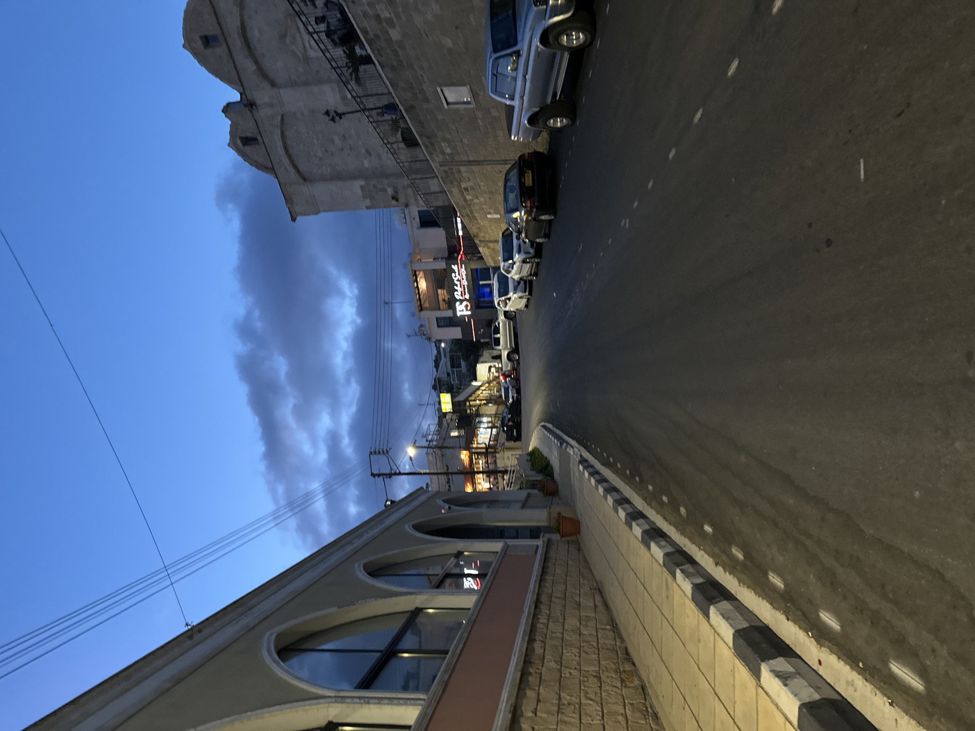 The image depicts a street scene in Peyia, Cyprus, during twilight. A paved road runs diagonally from the lower right to the upper left of the frame. The road has white dashed lines marking lanes and a black and white striped curb on the left side. To the left of the curb, a sidewalk is visible, constructed with light-colored paving stones. Adjacent to the sidewalk is a building with a pale yellow facade, featuring arched windows with dark frames. The building has a stone-like foundation.
On the right side of the road, a silver pickup truck is parked. Further down the street, several other vehicles are parked, including cars of various colors. Buildings line the right side of the street, some with illuminated signs, suggesting commercial establishments. One building has a domed roof and stone walls. The sky above is a deep blue with scattered dark clouds. Power lines stretch across the sky. A street lamp is visible on the right side of the road. The overall impression is of a quiet urban street as evening descends.