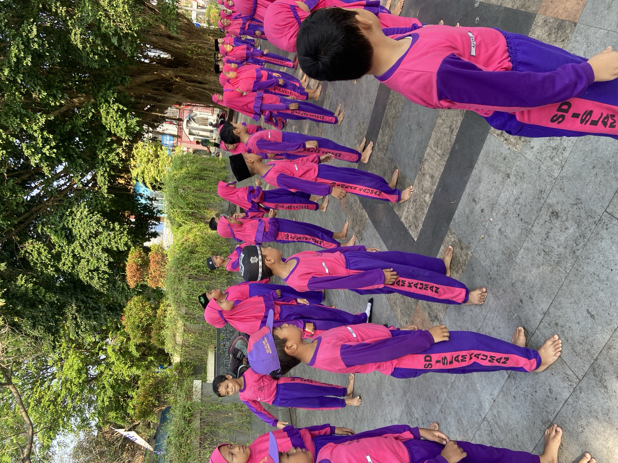 In front of a large, mature tree with sprawling roots and lush green foliage, a group of children, dressed in matching pink long-sleeved shirts and purple pants with a pink stripe down the side, are standing in formation on a paved outdoor area. The pants have "SD ISLAM KAUMAN" written vertically in pink lettering. Some of the children are wearing pink headscarves, and some are wearing black peci hats. They appear to be engaged in some sort of activity or lesson. In the background, there are more trees, some buildings with red accents, and what appears to be a white flag. The overall setting suggests a schoolyard or a public park in Kecamatan Pati, Indonesia. The lighting indicates it is daytime, likely morning or late afternoon, given the warm sunlight filtering through the trees.