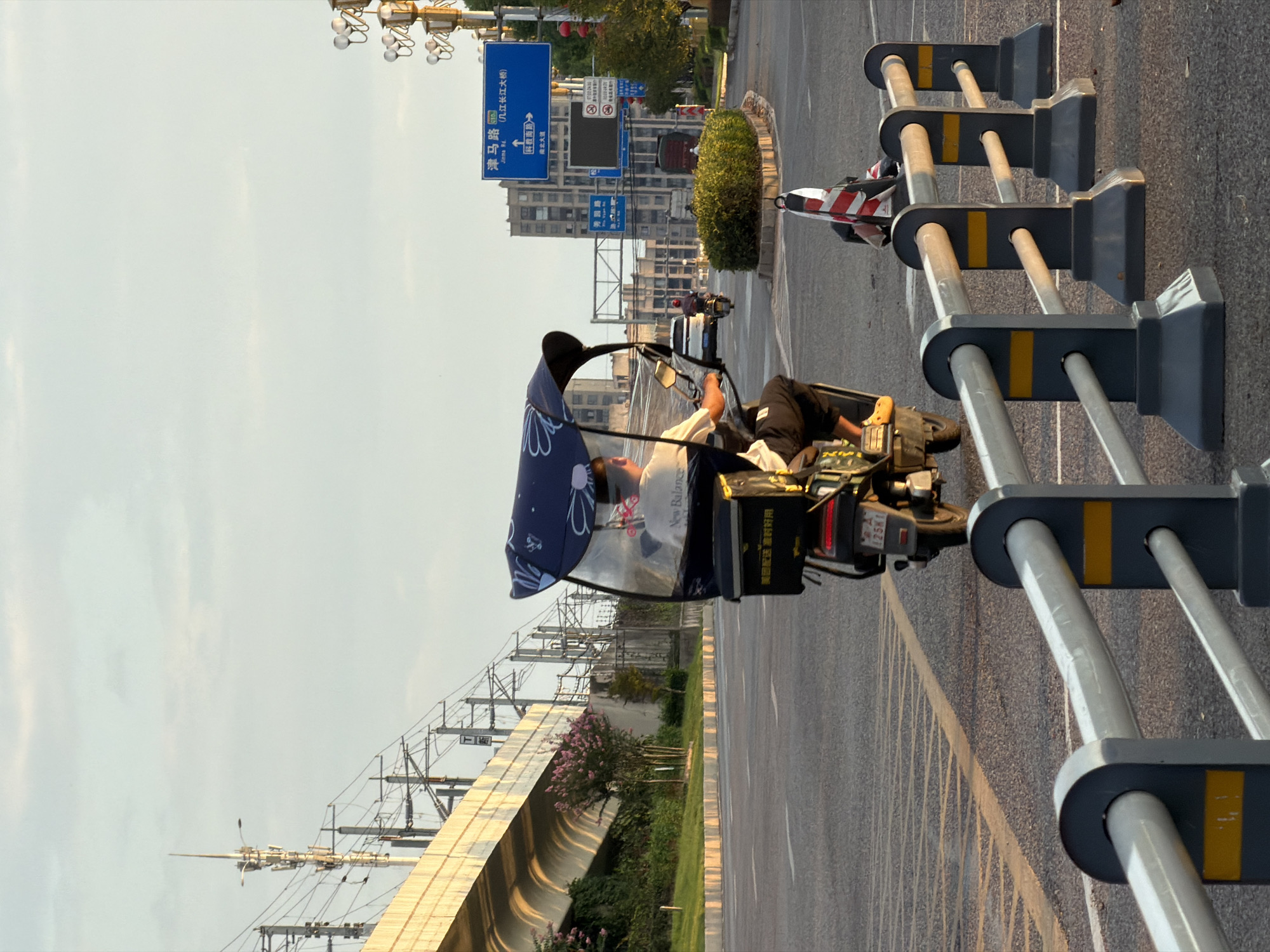 In Jiangjin, China, a person is riding a three-wheeled motorized scooter with a canopy on a paved road. The scooter has a black cargo box on the back. The rider is wearing a light-colored shirt with "New Balance" written on it in black and black pants. Yellow flip-flops are visible on their feet. The scooter has a license plate that reads "125KM." In the background, a large elevated structure, possibly a bridge or overpass, is visible, with overhead power lines. Buildings and streetlights are also present. Traffic signs, including one in blue with Chinese characters, are on the right side of the road. Red lanterns are hanging from poles on the right. The sky is overcast with a soft, diffused light, suggesting it is either early morning or late afternoon. The road is marked with white and yellow lines. A barrier with gray posts and silver rails, marked with yellow rectangles, lines the right side of the road.