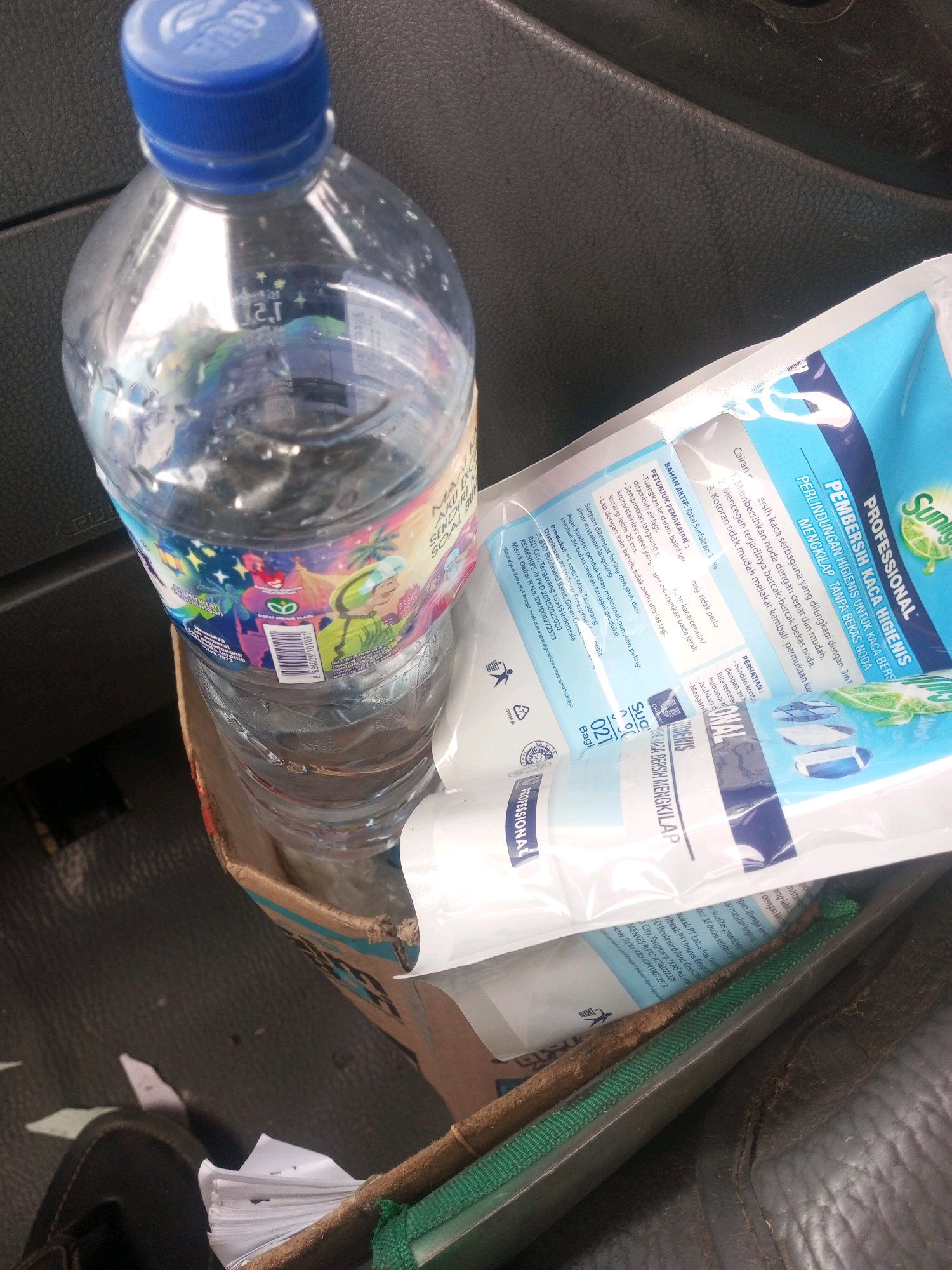 The image shows a plastic bottle of water with a blue cap sitting in a cardboard box. Next to the water bottle is a package of cleaning wipes with blue and white packaging. The scene appears to be in a vehicle, possibly a car or truck, as indicated by the dark, textured surface of the dashboard or interior. The lighting suggests it is daytime, possibly indoors or in a shaded outdoor area. The image is taken from a high angle, looking down at the objects. There are no people visible in the image. The overall impression is of everyday items stored within a vehicle.