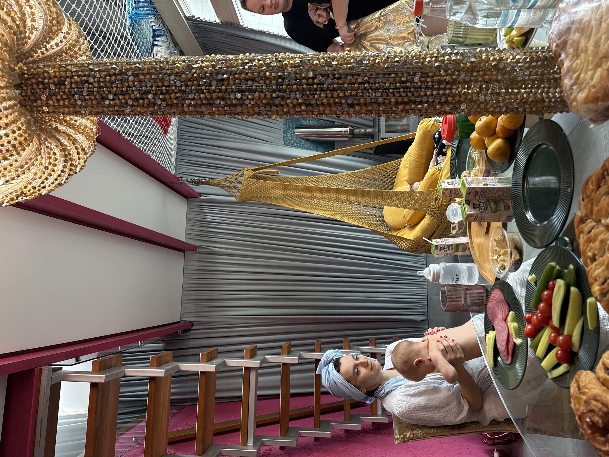 A rotated indoor shot depicts a woman with a light blue head covering, holding a baby. They are seated at a glass table laden with food items, including fruit, pastries, salami, and vegetables. Behind them is a silver-toned curtain. In the background, a hammock hangs, and a person is visible above it. Ornate decorations adorn the ceiling. The location is not specified to be in the city of Чёрное, Russia, and I cannot ascertain this.