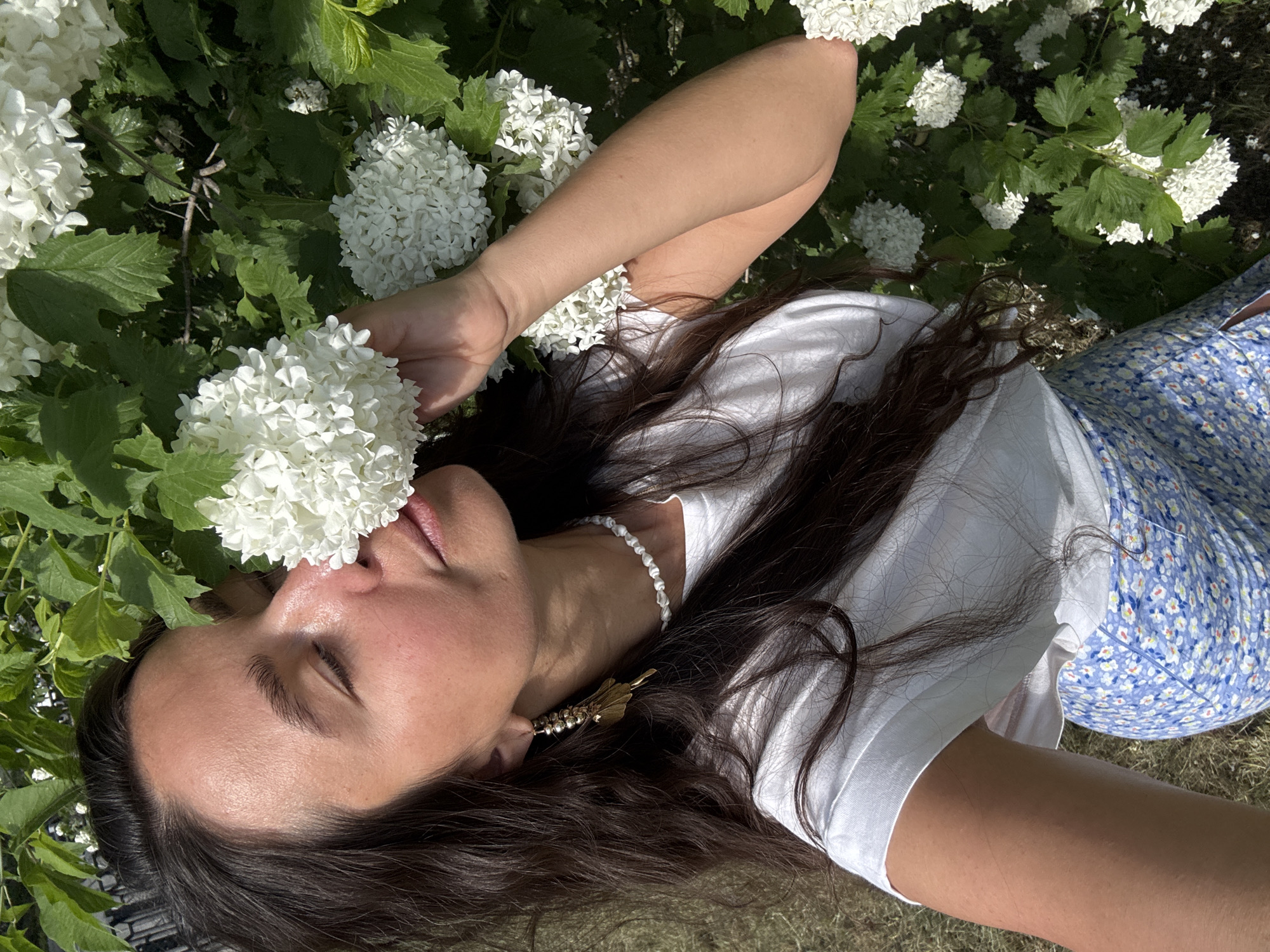 A woman lies on her back in a grassy area with white flowering shrubs. She is wearing a white t-shirt, a blue floral skirt, and a white beaded necklace. Her dark hair is spread around her head, and she has a gold earring. Her eyes are closed, and she is holding a white flower cluster near her face. The location appears to be an outdoor garden or park. The city and country are undetermined, despite the prompt's request.