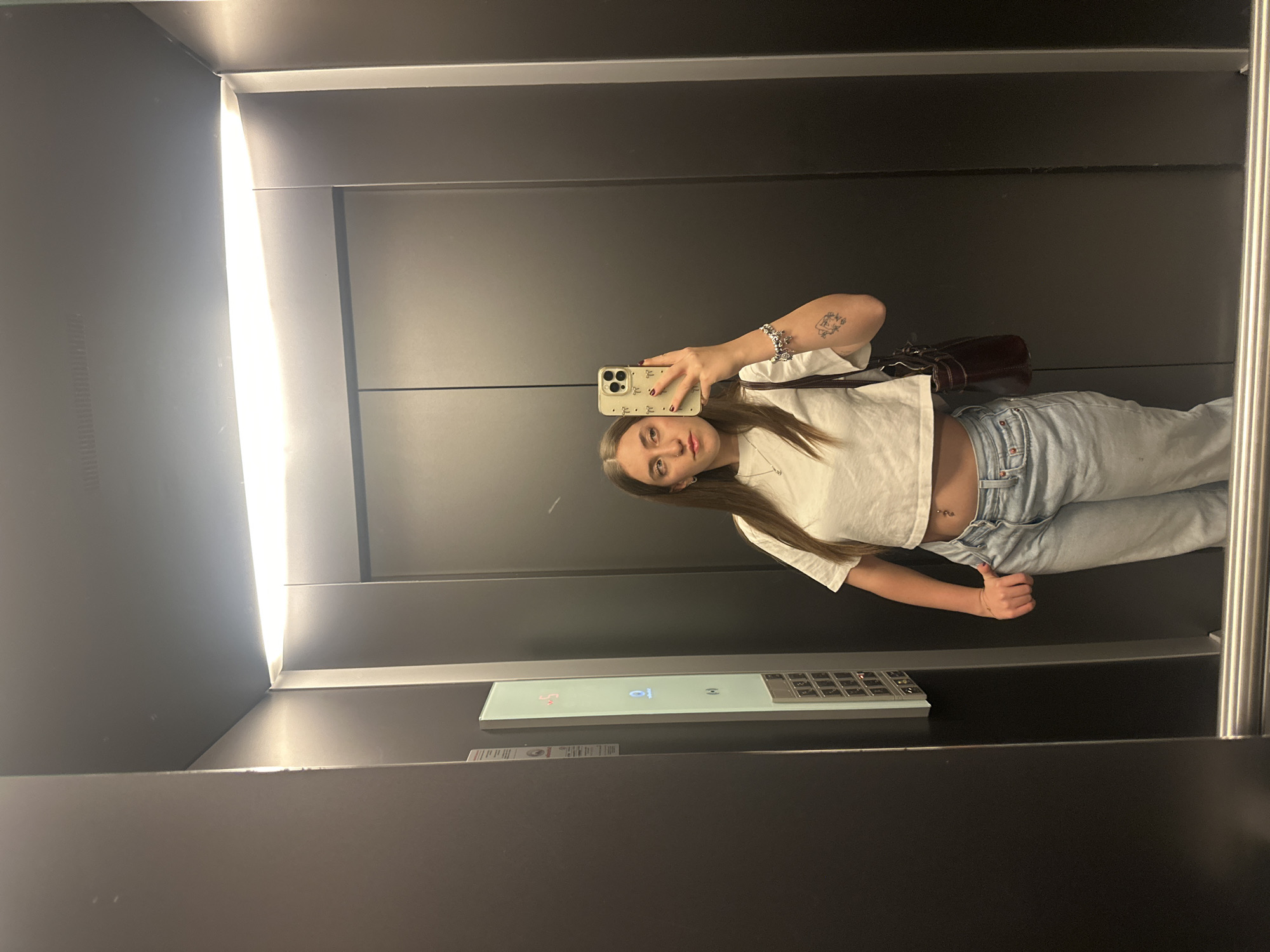 A woman is taking a selfie in an elevator with modern interior. She is wearing a white crop top, light blue jeans, and a brown shoulder bag. She is holding her phone in her right hand, which has a light-colored case with small designs. Her left arm has a tattoo. The elevator has a control panel with buttons and digital display showing the number 8. The elevator interior is dark, with a light panel illuminating one side. The location is likely in Huixquilucan de Degollado, Mexico.