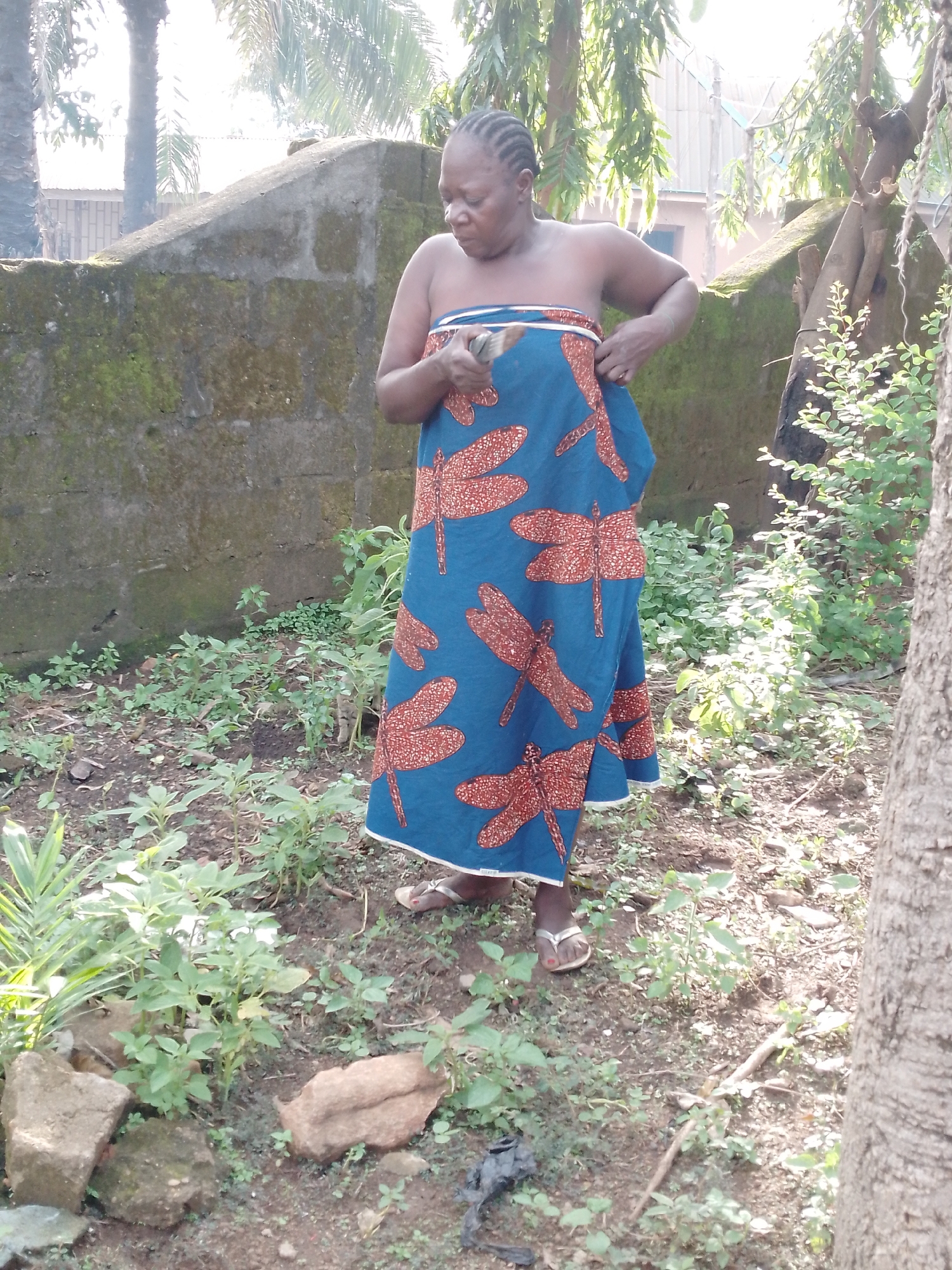 A full-body, medium shot, captured from a slightly low angle, shows an African woman standing outdoors. She has braided dark hair and is looking down at something she is holding in her hands. She is wearing a strapless blue dress with a pattern of orange dragonflies. Her bare shoulders and upper chest are visible. She is standing on a dirt ground with scattered green plants and some rocks. Behind her is a moss-covered, concrete wall. Trees and foliage are visible in the background. The lighting suggests it is daytime, with sunlight filtering through the leaves.