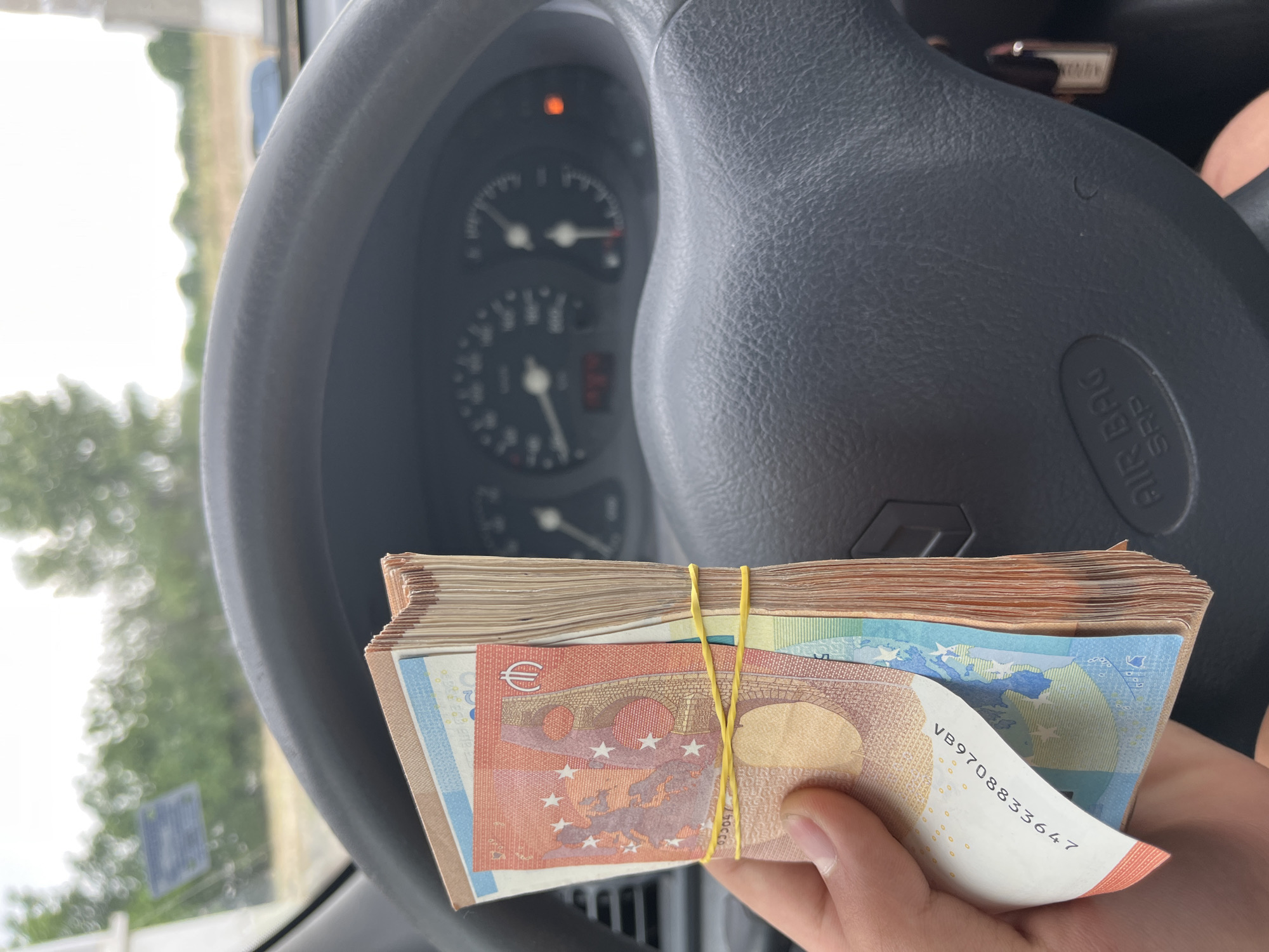The image captures a first-person perspective from inside a car, likely in Ivailo, Bulgaria, during daytime. The primary subject is a human hand holding a substantial stack of Euro banknotes, secured by a yellow rubber band. The top banknote is a 50 Euro note, recognizable by its reddish-orange color, architectural bridge design, stars, and the Euro symbol (€). A partial serial number, "VB9708833647", is visible on one of the notes.
The black steering wheel of the car dominates the foreground and midground, with the "AIR BAG SRP" inscription clearly visible on its lower right portion. Through the steering wheel, the car's dashboard and instrument cluster are visible, albeit slightly out of focus. A speedometer and tachometer are present, and an orange warning light is illuminated on the dashboard. Beyond the car's interior, a blurry outdoor scene is visible through the windshield, showing green foliage and trees against a bright, possibly overcast, sky, suggesting a natural or rural setting. The activity depicted is simply the act of holding the money.