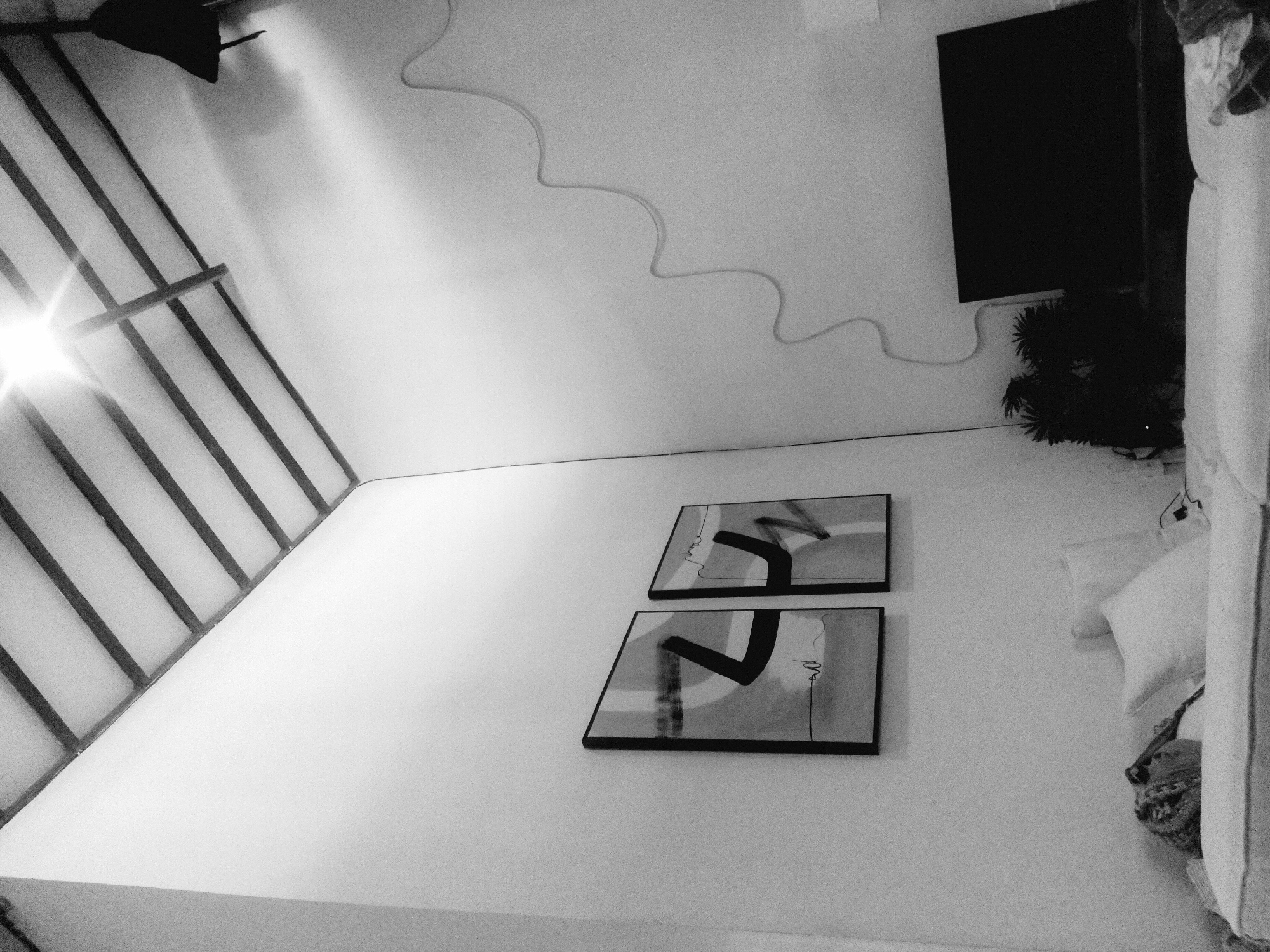 This is an interior shot, taken at an angle. On the left is a barred structure, likely a window. A light source is visible behind it. Two framed abstract art pieces hang on a wall. A wire runs across the upper part of the wall. To the right, a television is mounted. Below it are plants. A bed with pillows is situated to the right. This image was taken in North Kuta, Indonesia.
