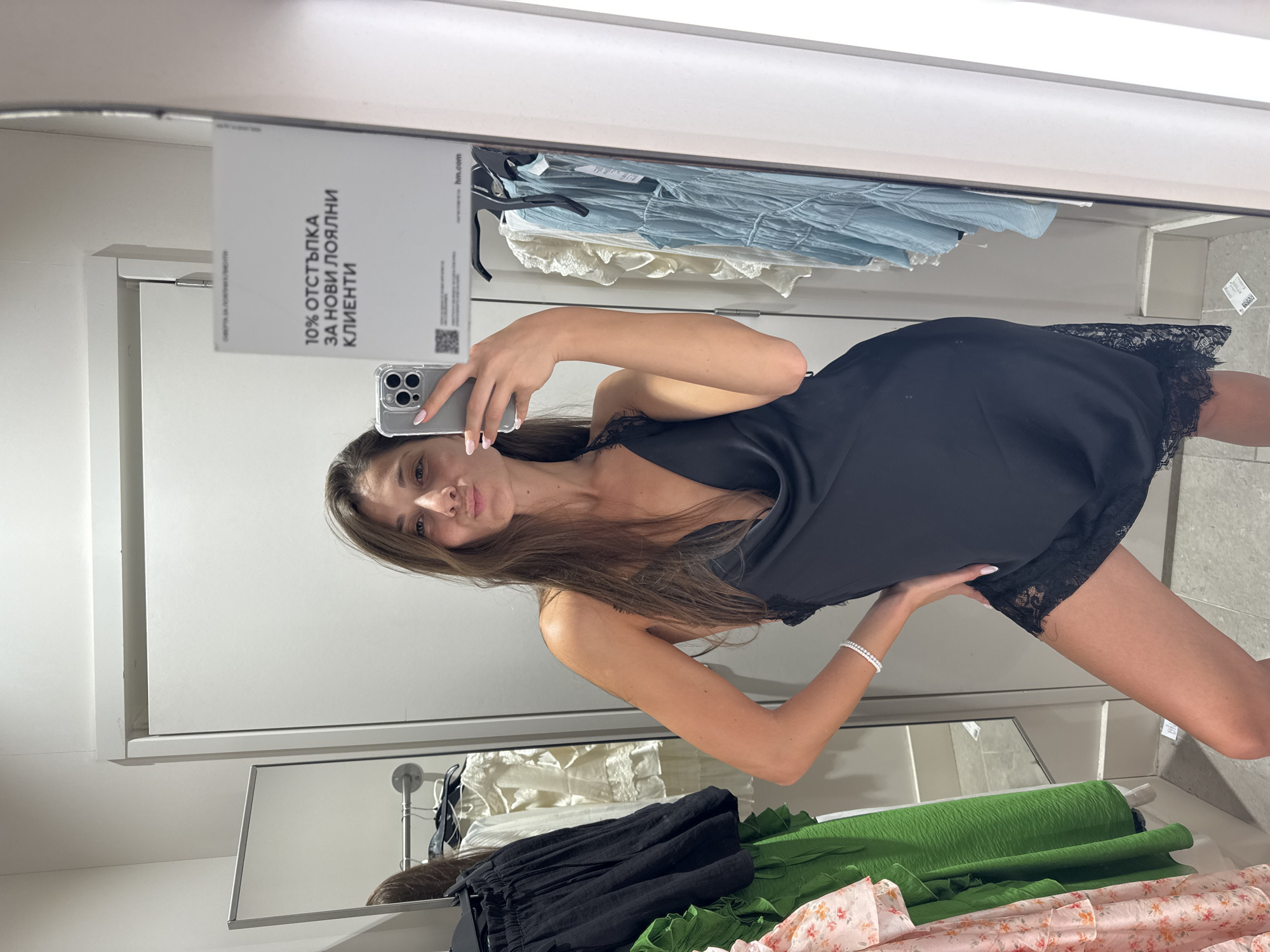 The image captures a young woman taking a mirror selfie in what appears to be a changing room or a retail clothing store located in Burgas, Bulgaria. The photograph is oriented sideways, with the subject and surroundings rotated 90 degrees clockwise.
The central subject is a woman with long, straight brown hair, wearing a black satin slip dress with delicate black lace detailing along the neckline and hem. She is holding a silver smartphone, likely an iPhone, in her right hand, positioned to capture her reflection. Her left hand is placed on her hip, and she looks directly into the phone's camera with a neutral, slightly pouting expression. She has light-colored nail polish and a multi-strand white bracelet on her left wrist.
The setting reveals elements typical of a clothing store. Behind the woman's reflection, a clothing rack holds various garments, including light blue and white textured fabrics. Below, on what appears to be a counter or lower shelf, are folded or rolled fabrics in black, vibrant green, and a pink floral pattern. The walls are white, and the area is brightly lit.
A prominent white sign is visible in the reflection, partially obscured by the woman and her phone. The text on the sign is in Cyrillic and reads: "10% ОТСТЪПКА ЗА НОВИ ЛОЯЛНИ КЛИЕНТИ", which translates to "10% DISCOUNT FOR NEW LOYAL CUSTOMERS". Below this, a QR code and the web address "hm.com" are also visible, suggesting the photo was taken inside an H&M store.