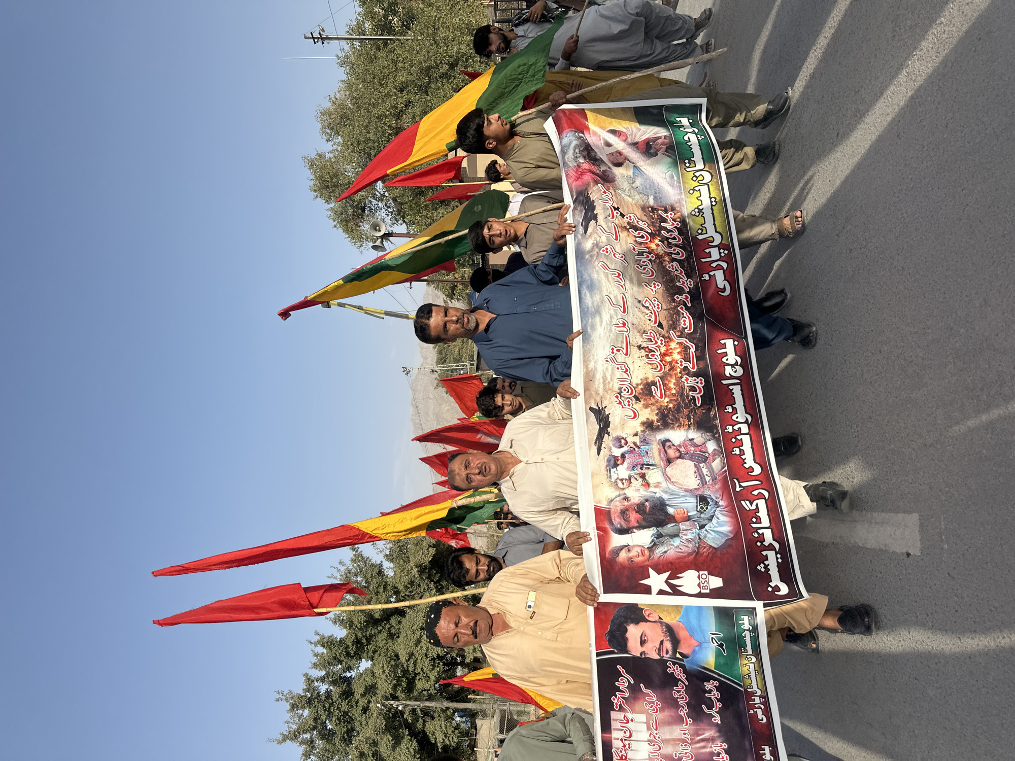 In Quetta City Tehsil, Pakistan, a group of men are participating in a protest or procession, holding a large banner and several flags. The banner, prominently displayed, features images of individuals, a military jet, and text in Urdu script. The flags are predominantly red, yellow, and green, hoisted on poles. The people are dressed in traditional South Asian clothing. They are walking on a paved road, with trees and buildings visible in the background. The sky is clear and blue, suggesting it is daytime. The overall scene depicts a public gathering, likely a demonstration or rally.
