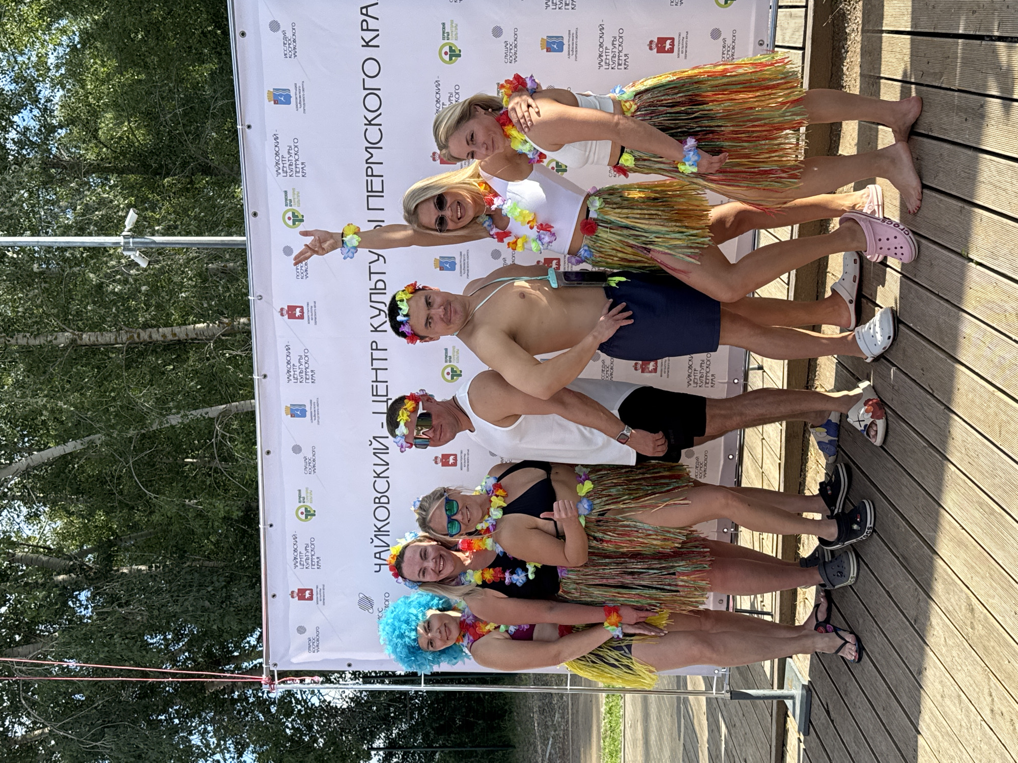 A group of five people, four women and one man, are posing for a photo in front of a banner. The people are dressed in a festive, tropical-themed attire, including colorful leis, flower crowns, and grass skirts. The woman in the front is wearing a bright blue wig. The man is topless, wearing sunglasses and a flower crown. The woman behind him is also wearing a flower crown and sunglasses, with a leis around her neck. The third woman from the front is smiling and wearing a leis. The woman in the back is also wearing a leis.
The banner behind them is white with text in Russian, which appears to be related to a cultural center in Chaikovsky, Perm Krai. There are also logos and graphics on the banner. The ground they are standing on is made of wooden planks, suggesting an outdoor setting. Sunlight is visible, indicating it is daytime, and the presence of trees in the background suggests a warm weather event. The overall mood seems joyful and celebratory. The text visible on the banner includes "ЧАЙКОВСКИЙ - ЦЕНТР КУЛЬТУРЫ ПЕРМСКОГО КРАЯ" which translates to "Chaikovsky - Center of Culture of Perm Krai". Other visible words include "ОТКРОЙ" (Open), "ИССЛЕДУЙ" (Explore), "СЛУШАЙ" (Listen), and "ОПРОБУЙ" (Try). The shoes worn by some of the people, particularly the pink "Crocs," are also a notable detail.
