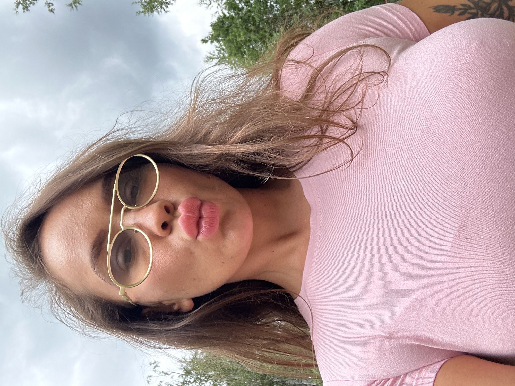 A woman with brown hair and glasses is captured in a close-up selfie outdoors. She is wearing a pink t-shirt and has her lips pursed for a kiss. The background consists of trees and a cloudy sky, suggesting a natural or park-like setting. There is no indication of the specific location within Duvankul', Russia.