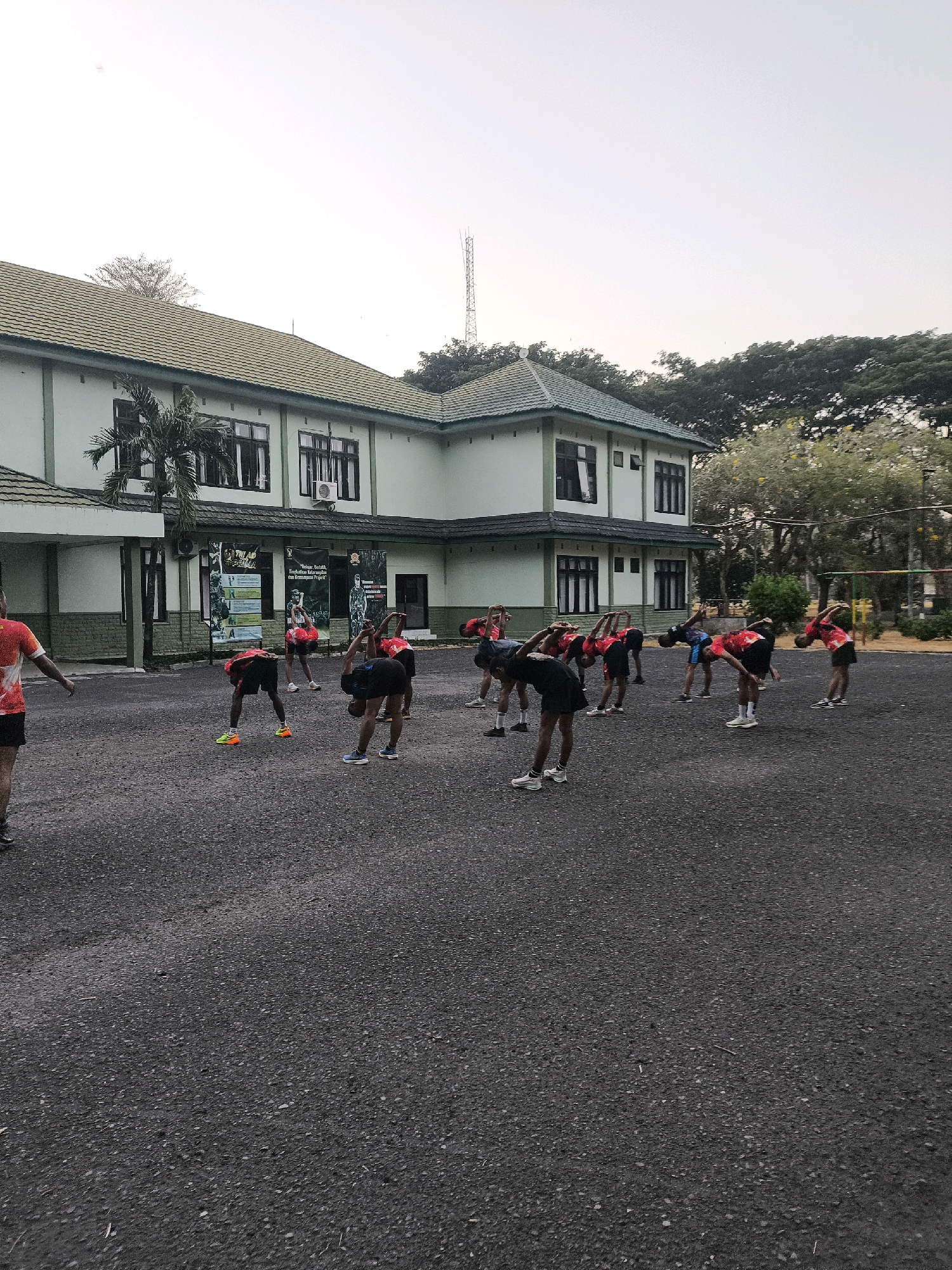 A group of individuals are performing stretching exercises in an outdoor paved area. The individuals are wearing athletic attire, predominantly red t-shirts and black shorts. They are in various stages of a forward bend, with hands reaching towards their feet or knees, and their backs arched.
The scene is set in front of a two-story building with a green tiled roof and light green walls. The building has multiple windows with dark frames. A palm tree is situated to the left of the building. Several large banners with text and images are displayed on the wall of the building.
The ground is covered in dark asphalt with visible small stones and some debris. In the background, trees and foliage are present, along with a tall communication tower and what appears to be a sports net structure. The sky is overcast and pale. The location is identified as unknown, Indonesia.