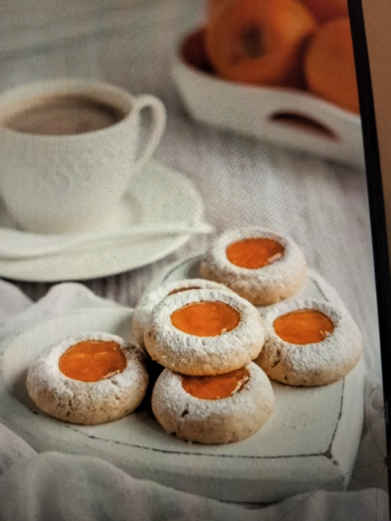 The image displays a close-up view of several thumbprint cookies filled with orange jam, dusted with powdered sugar, and arranged on a white plate. In the background, a white teacup filled with a dark beverage, likely coffee or hot chocolate, sits on a saucer. The setting appears to be a food display or a home kitchen. There is no indication of a specific city or location like Payagaboh, Indonesia. The image is devoid of any people, actions, or interactions. The environment is soft and blurred, with a focus on the cookies and the beverage.