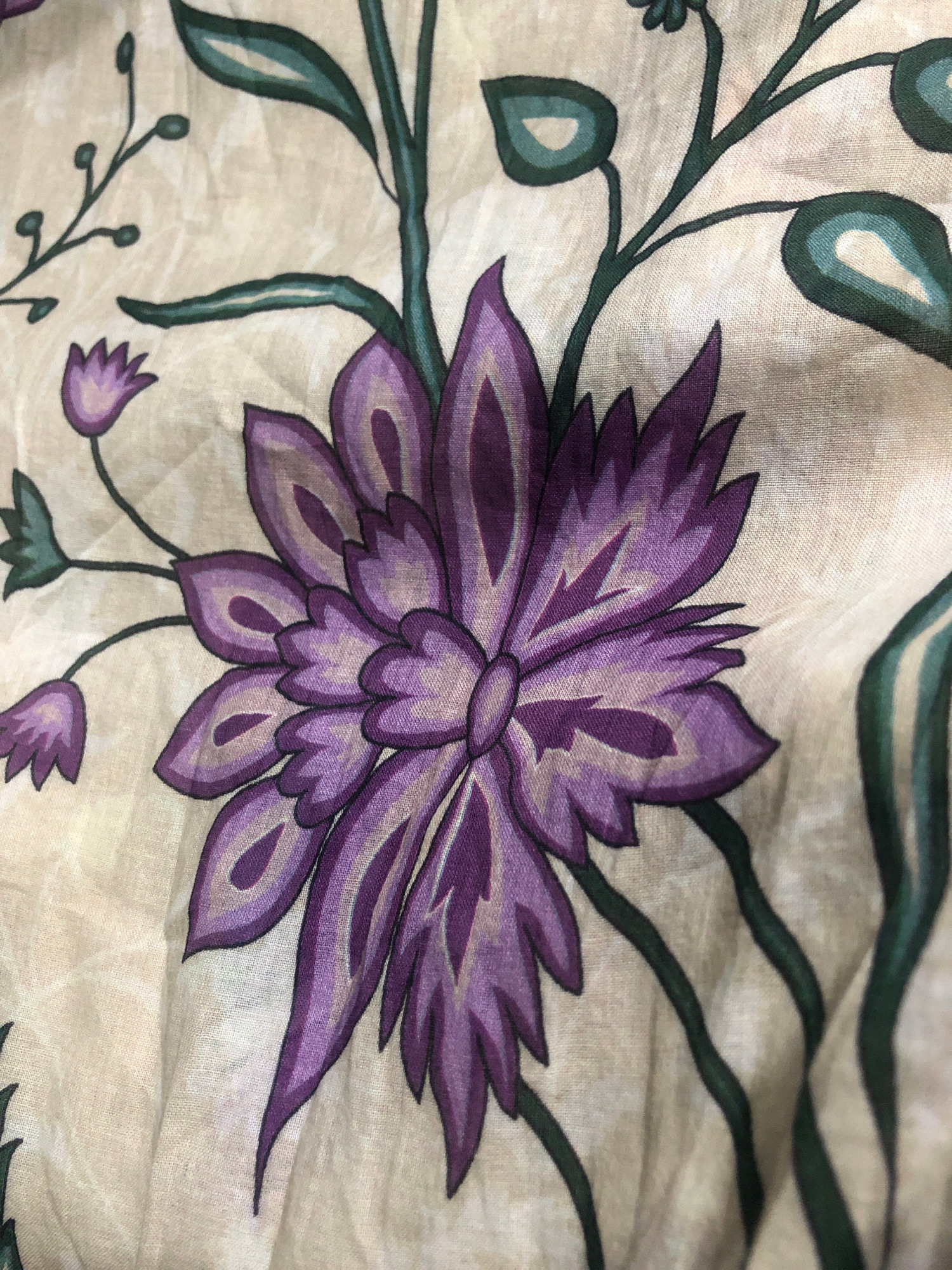 The image shows a close-up of a patterned fabric. The pattern features stylized floral elements, primarily in shades of purple and green, against a light beige background. A large, prominent purple flower with layered petals dominates the center of the frame. Smaller, bud-like purple flowers and green leaves and stems are scattered around the main flower. The fabric appears to be made of a lightweight material, possibly cotton or linen, and shows some wrinkles and folds, suggesting it might be draped or part of a garment or upholstery. The style of the pattern is reminiscent of traditional textile designs. There is no indication of a specific location in Karachi Division, Pakistan, within the image itself.