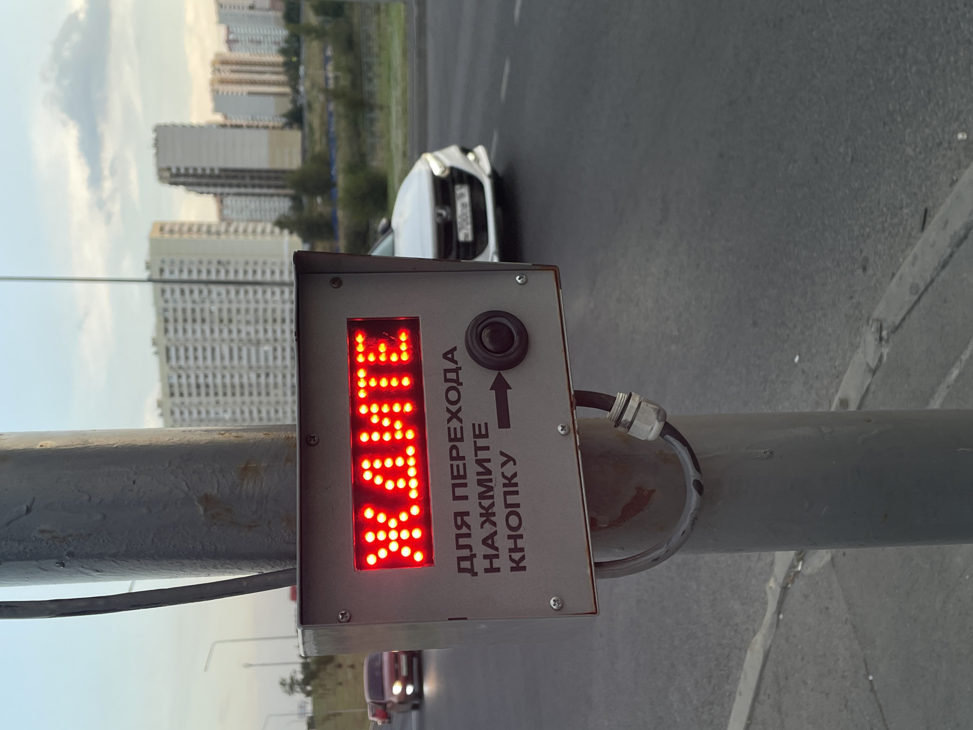 The image shows a close-up of a pedestrian crossing signal pole on a street in Rostov-on-Don, Russia. The signal has a red LED display showing the word "ЖДИТЕ" (WAIT) in large block letters, and below it, text reads "ДЛЯ ПЕРЕХОДА НАЖМИТЕ КНОПКУ" (TO CROSS, PRESS THE BUTTON) with an arrow pointing to a black button. The pole is grey metal, and a black cable is attached to a connector at the bottom of the signal box. In the background, there are cars on the road, residential buildings, and a cloudy sky. The overall setting appears to be an urban environment during the day.