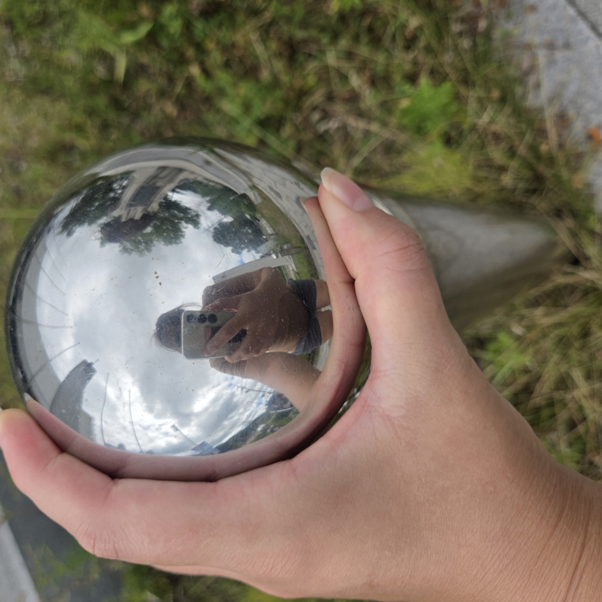 A hand is holding a highly reflective spherical object, likely a gazing ball, with a background of grass and pavement. Reflected in the sphere is the photographer, a person wearing a brown top and dark shorts, holding a mobile phone up to take a selfie. The reflection also shows a cloudy sky, trees, and a building with architectural details. The scene appears to be outdoors, possibly in a park or garden area. The location is identified as Dunnae-myeon, South Korea.