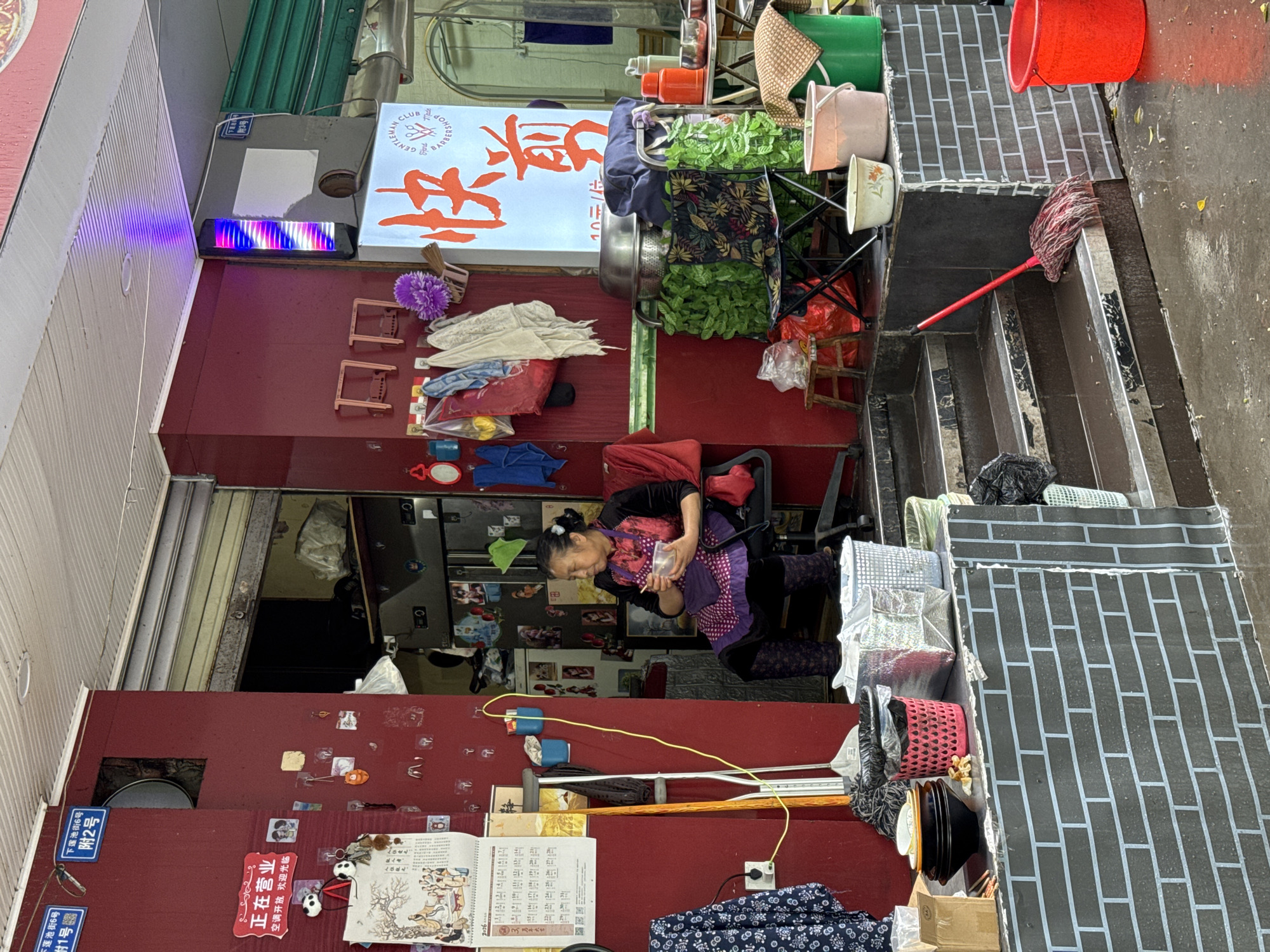 The image captures a street-level view in Jinjiang District, China. An older woman is seated on a chair within a shop or business establishment, seemingly engaged in a personal activity. Various objects and goods are scattered around, including bags, clothing, a calendar, and buckets. A red mop leans against a wall that appears to be constructed of gray bricks. A sign with Chinese characters is visible above, along with a barber shop sign. The environment looks cluttered, with a mix of commercial and personal items filling the space.