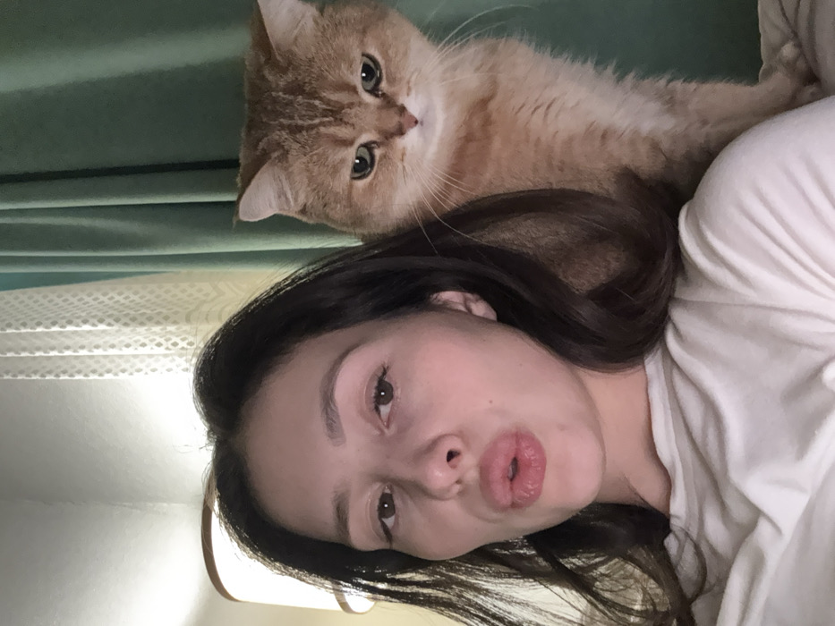 The image displays a close-up, indoor scene featuring one female individual and one domestic cat. The female, positioned on the left side of the frame, has dark hair and is observed from the chest up, wearing a light-colored, possibly white, top. Her head is angled slightly, and she is making a "kiss face" with her lips pursed. To her right and slightly behind her, a light brown/golden-furred cat, possibly a British Shorthair, is partially visible, looking forward with a neutral to slightly serious expression. In the upper left background, a lit lamp with a beige cylindrical shade emits a warm light. The background also includes two types of curtains: a sheer, patterned, light-colored curtain on the left, and a solid, dark teal curtain on the right. The specific location within the city of Yekaterinburg, Russia, cannot be determined from the visual information present in the image.