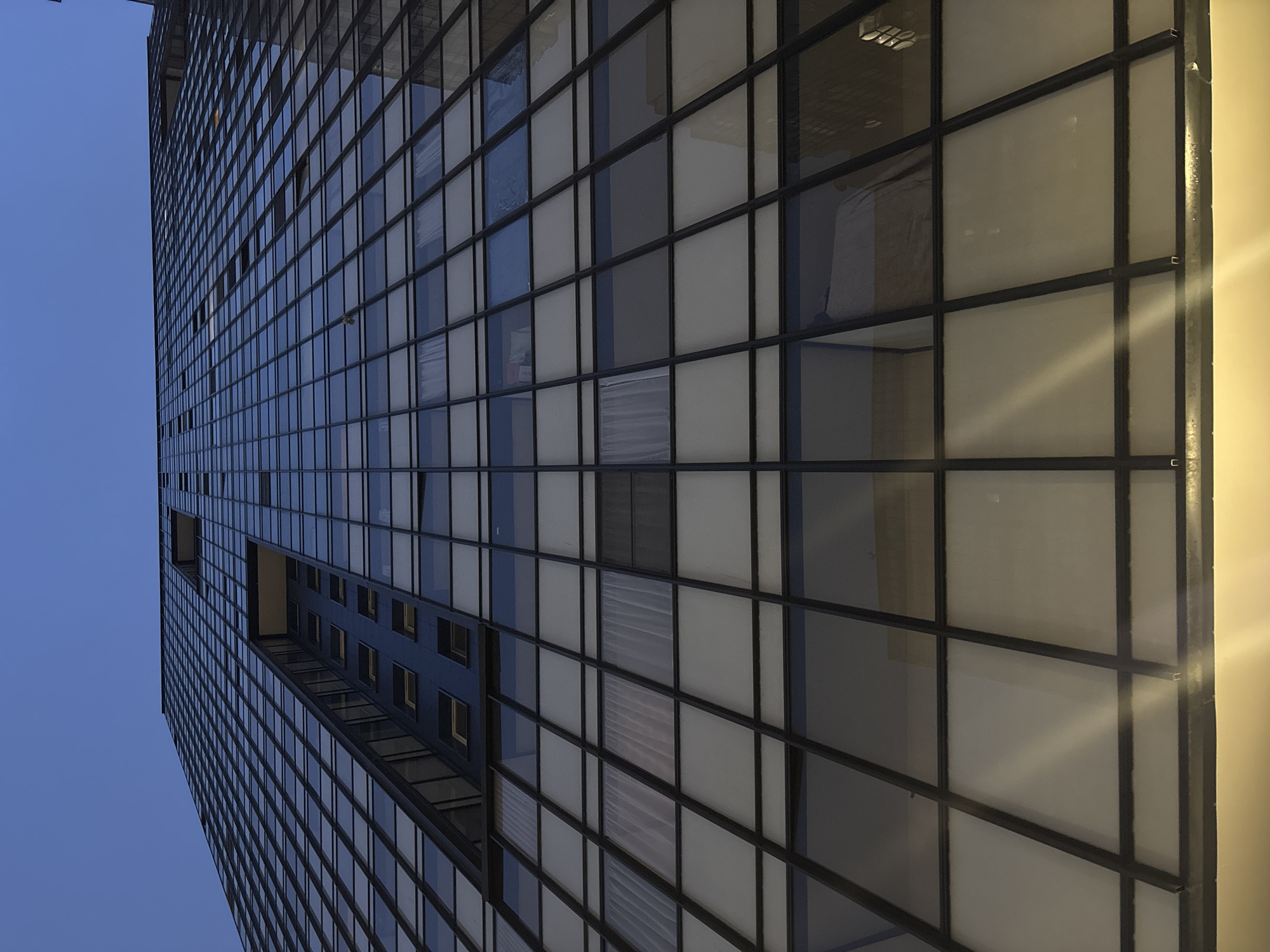 The image shows a tall, modern building with a grid-like facade. The facade is made of windows and dark metal frames, with a blue sky visible on the left side. There are some apparent details in the windows, such as some blinds or curtains. It is hard to pinpoint the exact location in Unknown, Unknown.