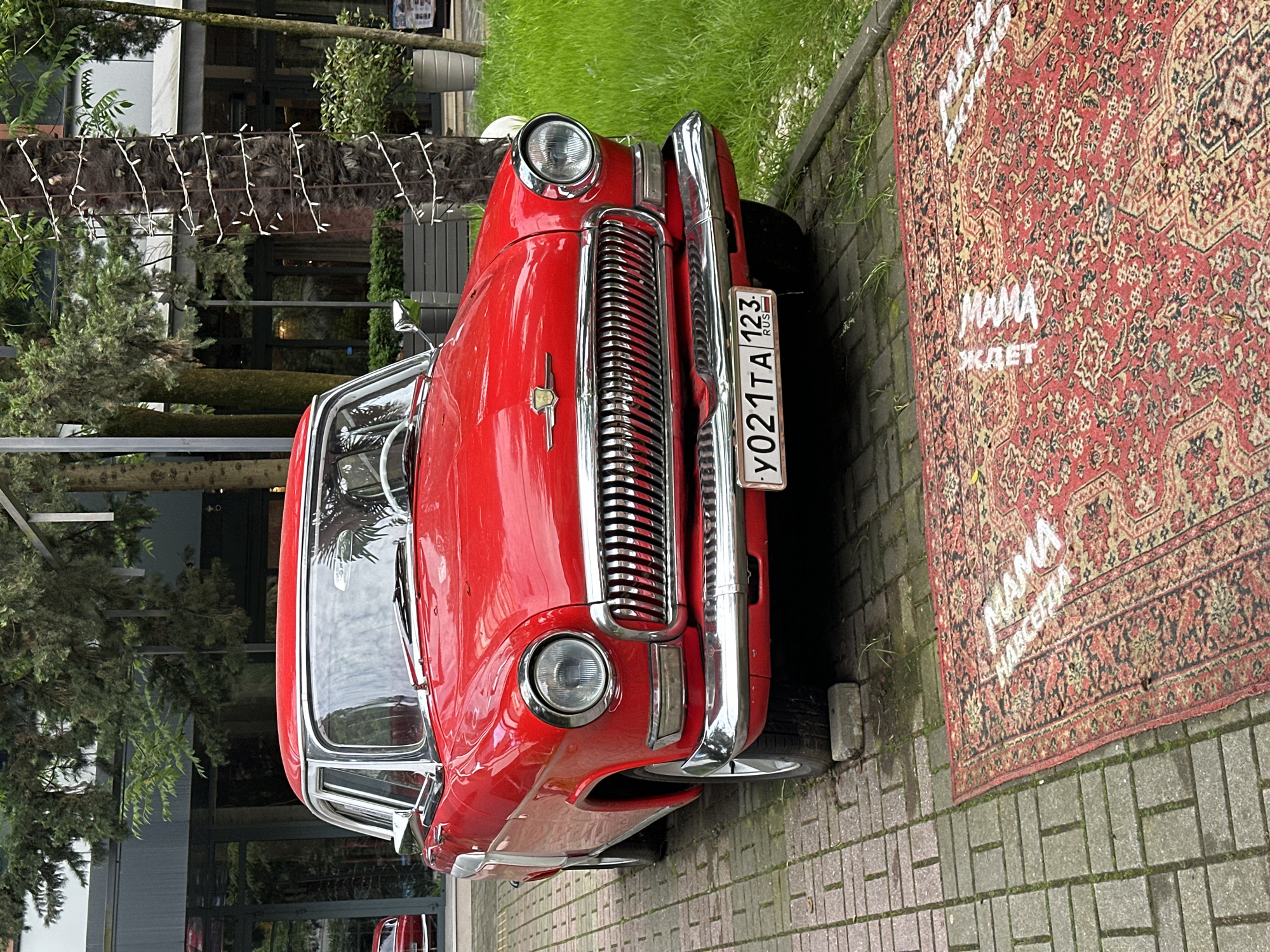 The image captures a vintage red car parked on a brick-paved area. The car is a classic model, possibly a GAZ-21 Volga, recognizable by its prominent chrome grille and round headlights. The license plate indicates it is registered in Russia. Next to the car is a traditional red carpet with text in Russian, seemingly painted onto it. The backdrop features greenery and a building facade, suggesting an urban setting. Given the license plate and architectural style, the location is likely in the city of Sochi, Russia. There are no people visible in the immediate scene. The interaction between the car and carpet suggests a deliberate arrangement for display or decoration.
