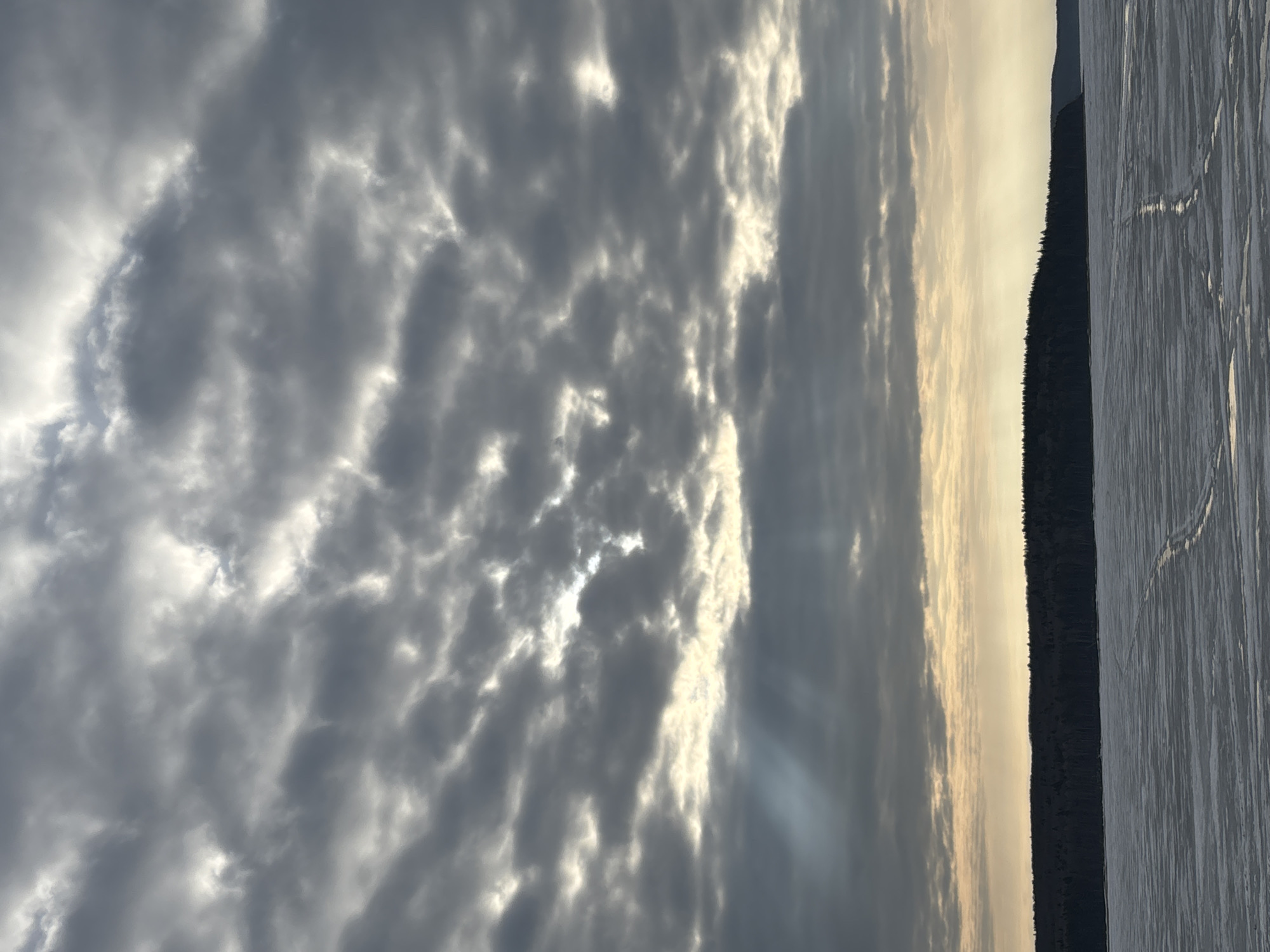 The image depicts a landscape with a cloudy sky dominating the upper portion. The clouds are layered and appear to be illuminated from behind, with some rays of light breaking through. In the midground, there is a dark band of trees, possibly a forest or a line of trees along a horizon. The foreground shows a frozen expanse, likely a lake or a large body of water covered in snow or ice, with some visible tracks on its surface. The location is potentially Chaikovsky, Russia, based on the user's prompt, but there are no specific indicators to confirm this. No people or actions are visible in the image. The environment suggests a cold, possibly winter, scene.