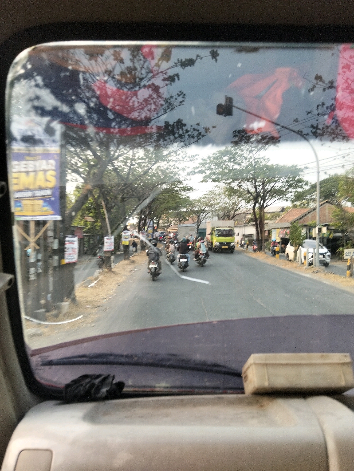 The image captures a street scene in Semarang, Indonesia, viewed from inside a vehicle. The foreground shows the interior of the vehicle, including the dashboard and a windshield wiper. The windshield itself is reflective, showing a distorted image of trees and a red fabric. The middle ground depicts a road with several motorbikes and a green truck. The road is lined with trees and buildings. A billboard with text in Indonesian, possibly advertising "GEBYAR EMAS," is visible on the left side of the road. The scene appears to be during daytime, with clear skies.