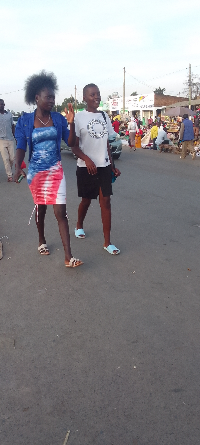 This full-shot street photograph, taken in Kakunga, Kenya, captures two women walking on an asphalt road in the foreground, with a bustling market scene behind them. The lighting suggests late afternoon or early morning.
The woman on the left has dark, curly hair and wears a blue jacket over a blue, white, and red tie-dye dress, accessorized with a necklace and light flat sandals. She holds a phone in her left hand and her right hand is raised, possibly in a wave or gesture. The woman on the right has short hair and is wearing a white t-shirt with "BURBERRY ENGLAND" printed on the front, a black skirt, and light blue slide sandals. She carries a dark shoulder bag and appears to be smiling or laughing, with her right hand also raised.
In the background, numerous people are engaged in market activities, with various goods displayed on the ground and makeshift stalls. Buildings line the street, bearing signs that include partially visible text such as "CYBER ALQUBA", "REPAIR...", "DAILY COSY", and "HARDWARE". A light-colored car is partially visible near the market area. Utility poles and overhead wires are present along the street. The sky is bright and clear, indicating good weather conditions.