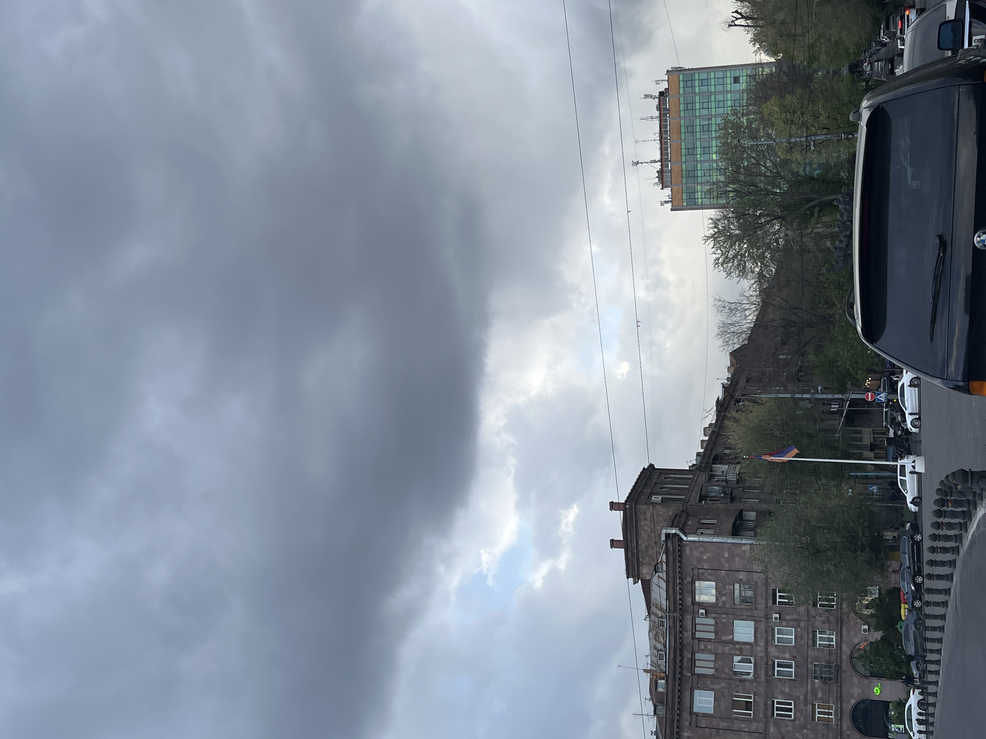 The image shows a street scene in Yerevan, Armenia, taken on a cloudy day. The left side of the frame is dominated by heavy, dark clouds. To the right are buildings, the closest of which are made of dark stone with multiple windows and chimneys. An Armenian flag is visible on a pole near one of the buildings. A modern, green-tinted glass building is partially visible in the background. The right side of the frame is occupied by a street with parked cars and traffic, including the rear of a black van in the foreground. Utility wires cross the sky.