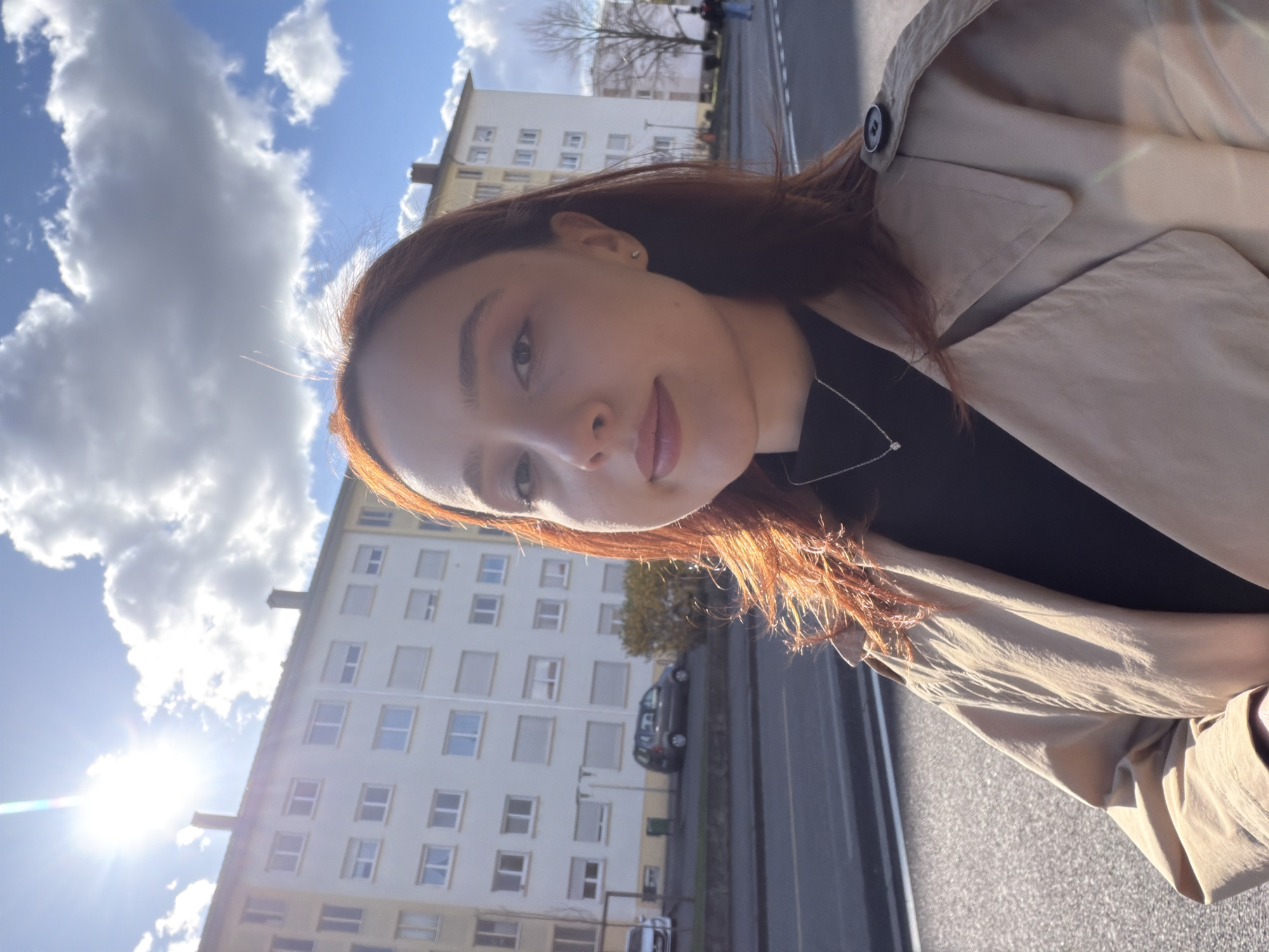 A front-facing outdoor photograph depicts a young adult female on a residential street in Strasbourg, France, under a bright, sunny sky with scattered clouds. The primary subject, positioned in the foreground and slightly to the right, has fair skin and medium-length reddish-brown hair backlit by the sun, creating lens flare in the upper left. She wears a black top, a light beige trench coat, and a silver necklace with a small pendant. She looks directly at the camera with a neutral expression. In the background, two multi-story apartment buildings, predominantly white or pale yellow, feature numerous rectangular windows, some with light-colored roller shutters, and multiple chimneys. A paved street with double solid white lines and a light-colored pedestrian crossing is visible in the mid-ground. Several vehicles are parked along the street, including a silver hatchback and a darker grey SUV/minivan. Sparse vegetation, including a low bush with yellowish leaves and a bare deciduous tree, lines the street. A single, indistinct figure is visible walking away on the far right side of the street near the bare tree.