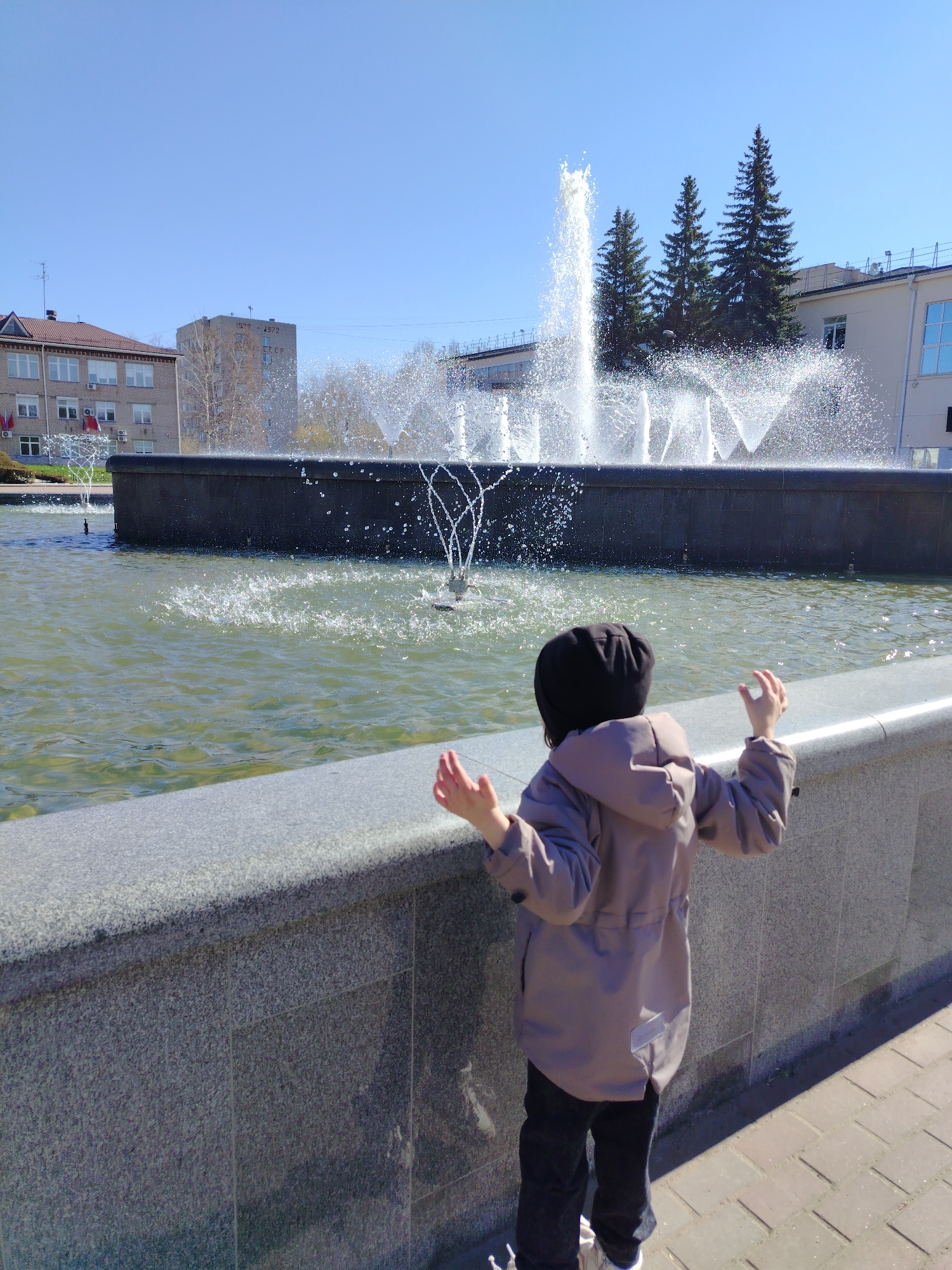 A child in a hooded jacket and dark pants stands at a fountain in Chaikovsky, Russia, facing the water. The fountain is a long, dark structure with multiple water jets spraying upward. Buildings are visible in the background, along with some trees. The day appears sunny. The child has their arms raised slightly, seemingly in reaction to the fountain. The surface in front of the fountain is paved.
