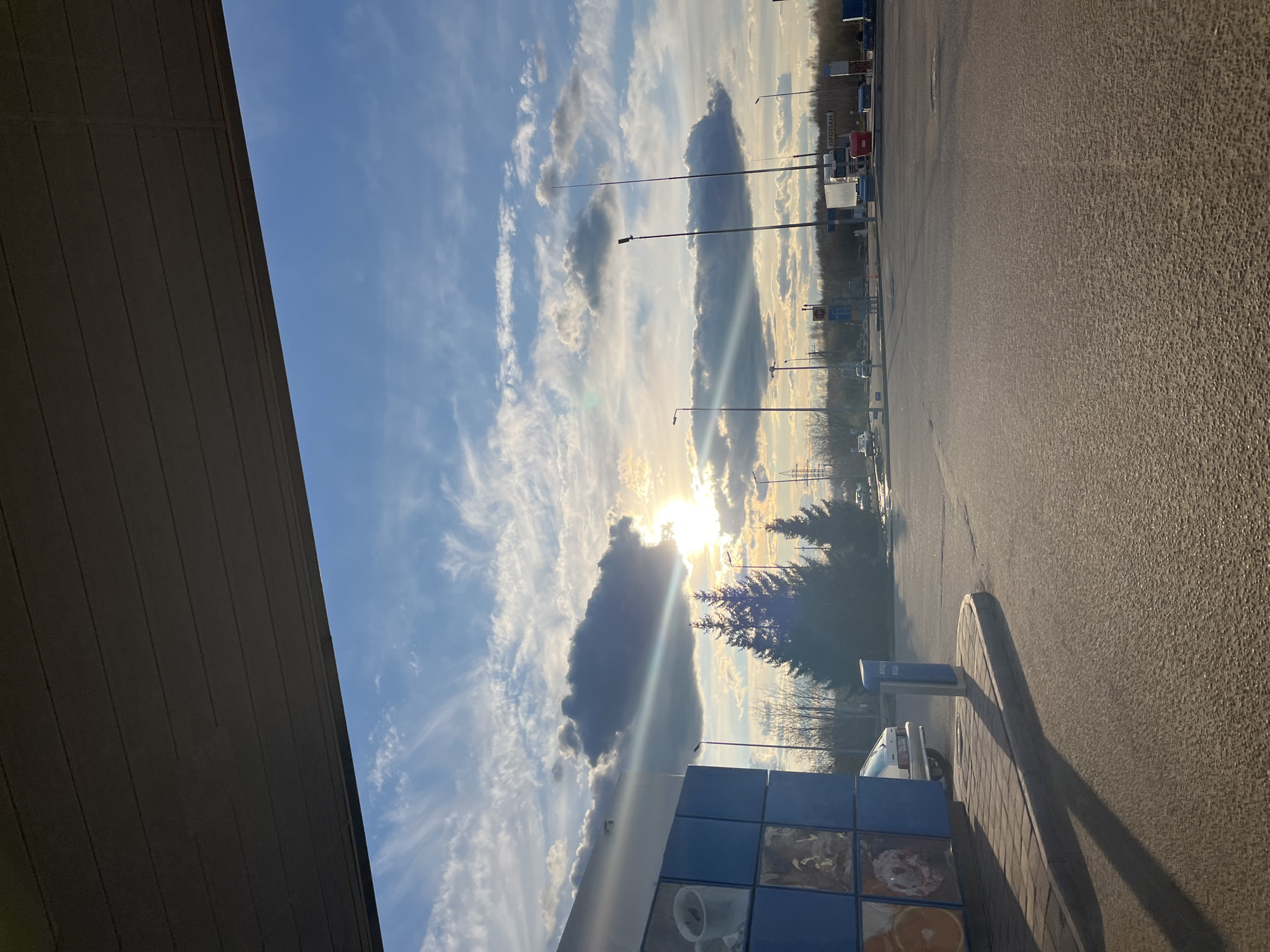 The image depicts an outdoor scene in Klin, Russia. The photograph is oriented with the horizon running vertically along the right edge. Overhead, the sky is bright blue with numerous wispy white clouds and a large, dark gray cloud obscuring the sun. Light rays emanate from behind the cloud. A row of streetlights lines the roadside. A flat, gray asphalt surface occupies the lower right portion of the frame. In the foreground, on the left side, part of a building is visible. It has a gray, horizontal paneled overhang and a lower section of blue tiled panels. A small part of a car is visible near the blue tiles. Trees are visible in the distance.