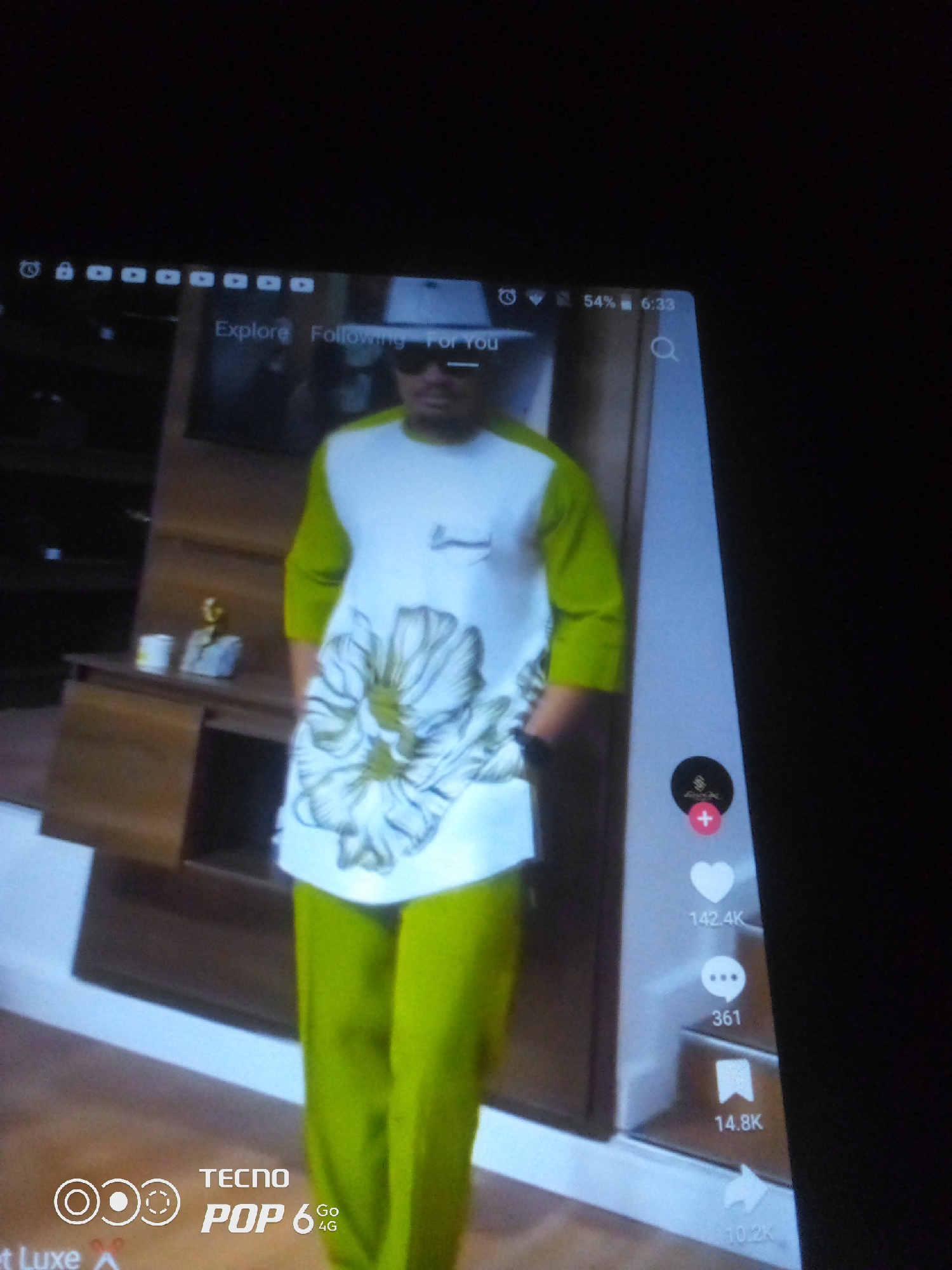 The image displays a screenshot of a mobile phone, likely a TikTok interface, showing a person in a green and white outfit with a floral pattern. The person is wearing sunglasses and appears to be standing indoors, possibly in a store or a home with wooden furniture visible in the background. The phone screen exhibits social media icons, including likes, comments, and shares, with associated numbers. The status bar at the top shows the time as 6:33 and battery level as 54%. The bottom left corner of the screen displays "TECNO POP 6 Go 4G". There is no information in the image that indicates the location is Howayi, Nigeria.