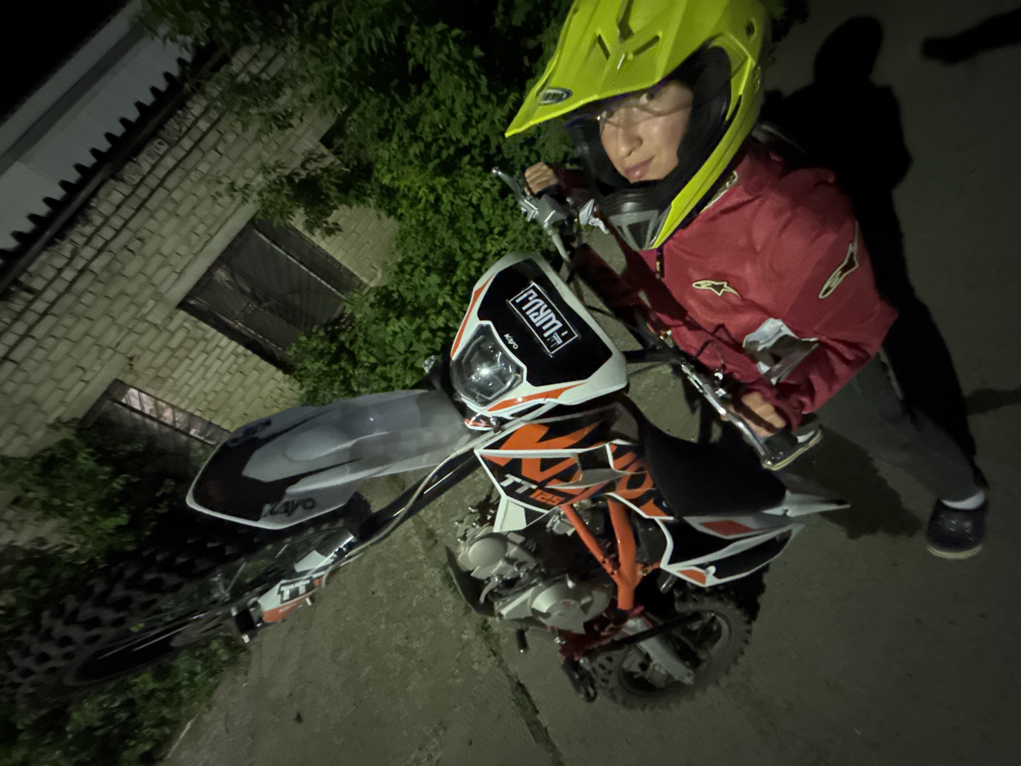 A person wearing a yellow motocross helmet, glasses, and a red jacket is sitting on a Kayo TT125 dirt bike. The bike is mostly white and orange with black and gray accents. The person is holding the handlebars. The location is an outdoor space at night in the city of Chaikovsky, Russia. There is a brick building with a grated window behind the rider. There is vegetation between the building and the rider. The ground appears to be a paved surface. The scene is illuminated by an external light source, creating shadows.