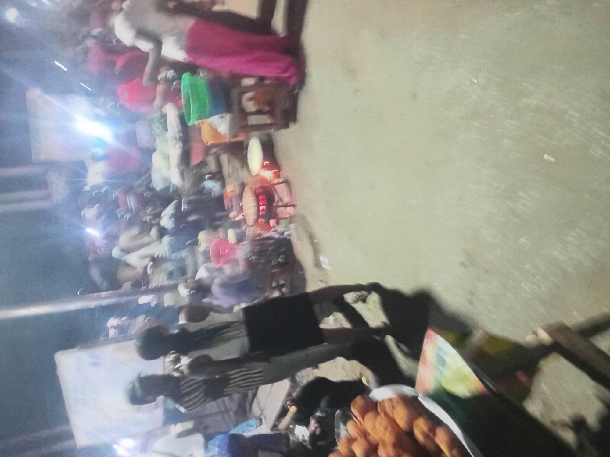 The image appears to be taken at a nighttime street market in Lokoja, Nigeria. Numerous people are present, possibly engaging in commerce around street vendors. Several people are wearing light-colored clothing and some are wearing pink clothing. Vendors' stands with various items, including what appears to be cooked food, are visible. The ground is asphalt. The lighting is a combination of streetlights and possibly vendor lighting.