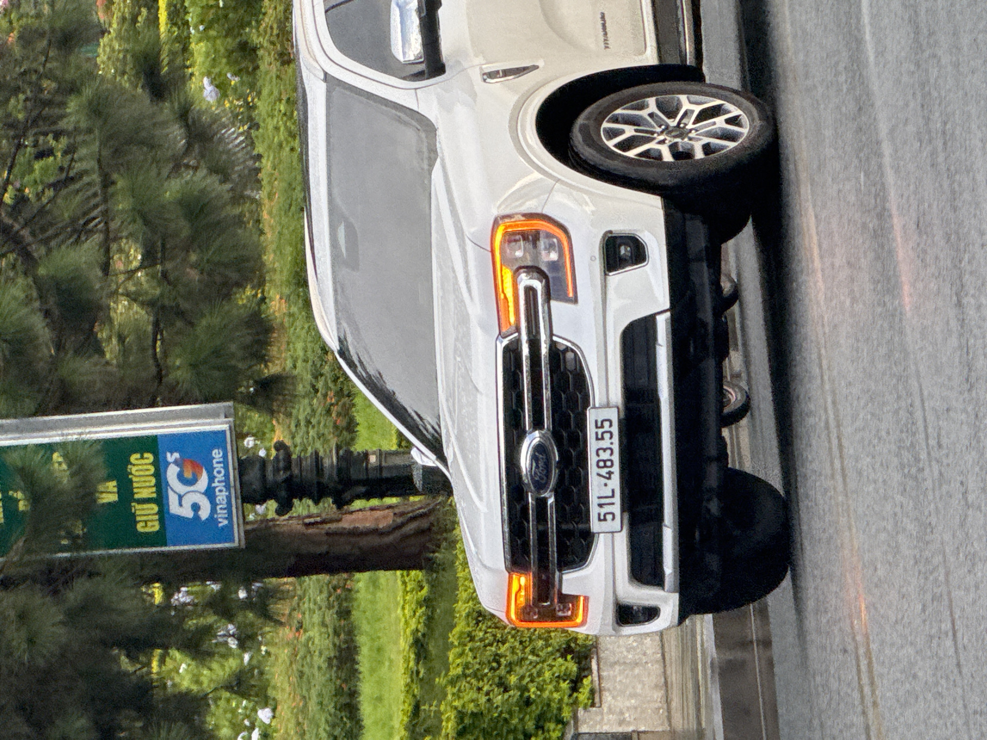 A white Ford Everest SUV is parked on a wet asphalt road in Da Lat, Vietnam. The SUV has its daytime running lights illuminated and its license plate reads "51L-483.55". In the background, there is a dense green hedge and trees. A dark, ornate lamppost with a green sign is visible to the left of the vehicle. The sign features yellow Vietnamese text and a blue box with a white "5G" logo and the Vinaphone branding. The environment suggests an overcast or rainy day due to the wet road and muted lighting.