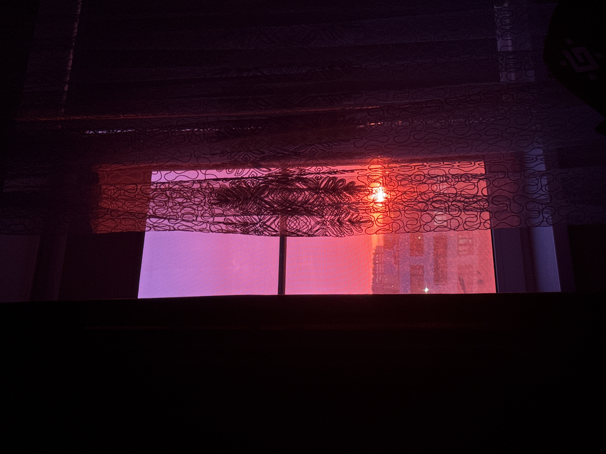 The image shows a view from inside a room, looking out a window with patterned curtains. The sunlight is intense, casting a red and orange glow through the curtains and onto the building visible outside. The lower part of the image is dark, obscuring the room's interior. No people or specific objects are discernible besides the window, curtains, and distant building. The location is Unknown, Unknown.