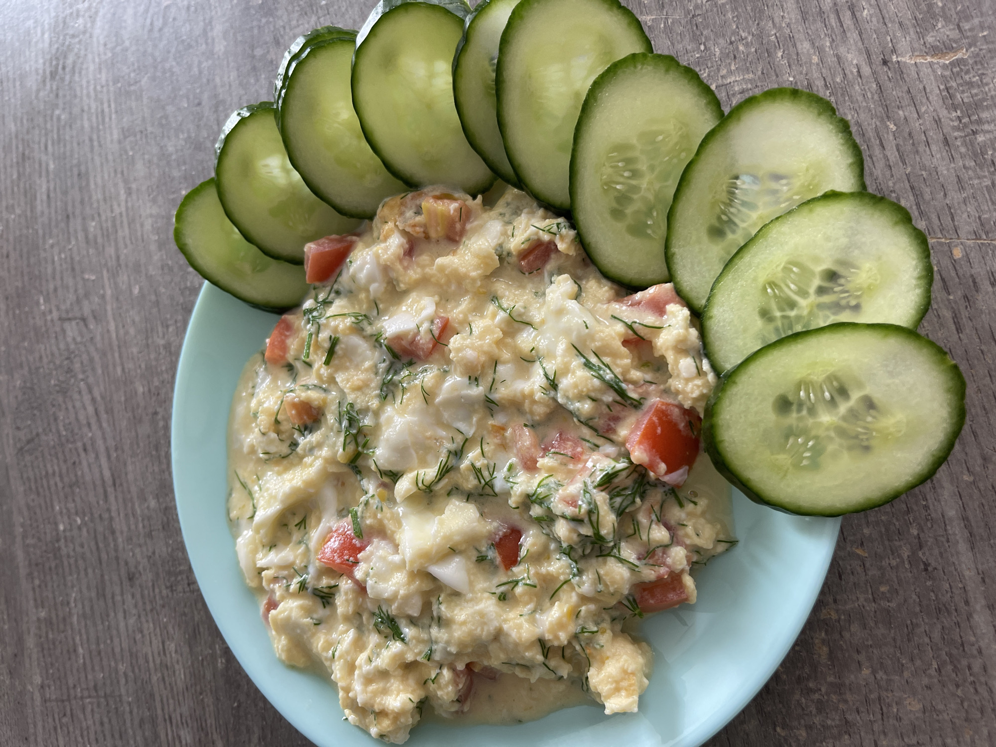 A close-up overhead shot captures a light blue plate of food, placed on a dark wooden table. The plate contains a serving of scrambled eggs with chopped tomatoes and dill. Sliced cucumbers are arranged in a semi-circle along the right side of the plate. The background is out of focus, but appears to be a wooden surface. No people are visible in the image. The image is a still life of food and is not specific to Blagoveshchensk, Russia.
