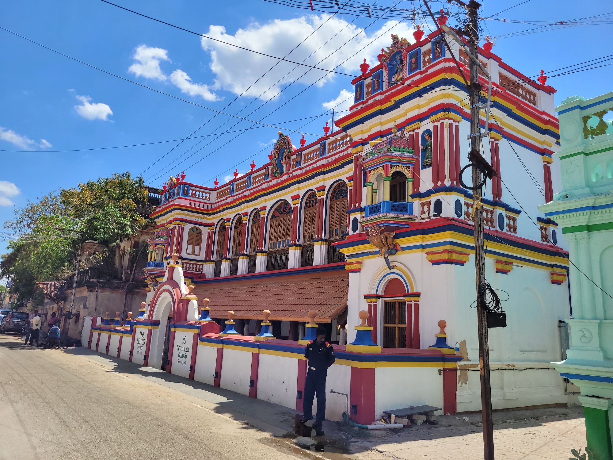 This image captures a vibrant street scene dominated by a grand, multi-storied building under a clear blue sky with scattered white clouds. The building is ornately decorated and painted in striking colors of red, yellow, blue, and white, with intricate architectural details, arches, balconies, and decorative figures along its roofline.
On the left side of the frame, a paved street stretches into the distance, where a few people are visible near parked vehicles. A person in a dark blue uniform stands near the front of the colorful building, close to a water spigot. The building's white compound wall features a decorative archway and is also painted with red, yellow, and blue stripes. Visible text on this wall reads "THE LOTUS PALACE CHETTINAD EST 1905" in English, along with corresponding text in Tamil script.
A tall utility pole, laden with numerous electrical wires, stands prominently in the foreground on the right, partially obscuring parts of the building. To the far right, a section of another building, painted in lighter green and blue tones, is partially visible. The scene is illuminated by bright daylight, casting clear shadows, indicating a sunny day. The architectural style suggests a South Indian setting, likely Chettinad, as indicated by the visible text.