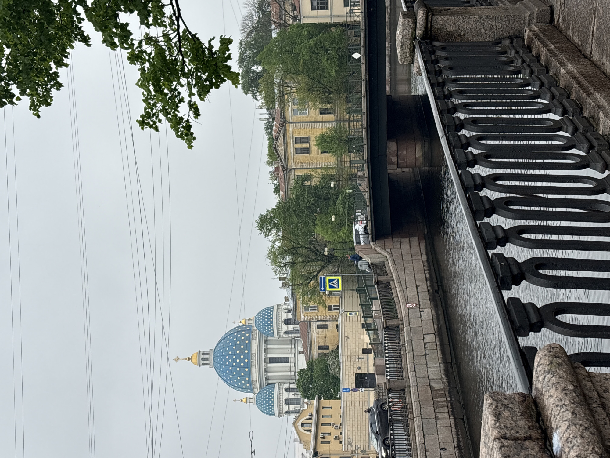 The image depicts a view of the Trinity Cathedral in Saint Petersburg, Russia, taken from a perspective that includes a canal and its embankment. The cathedral is recognizable by its blue domes dotted with stars. The canal is lined with a stone embankment and a metal railing. Overhead, there are several electrical wires running parallel to the top edge of the frame. Tree branches with green leaves appear in the upper left corner. The sky is overcast, suggesting a cloudy day.