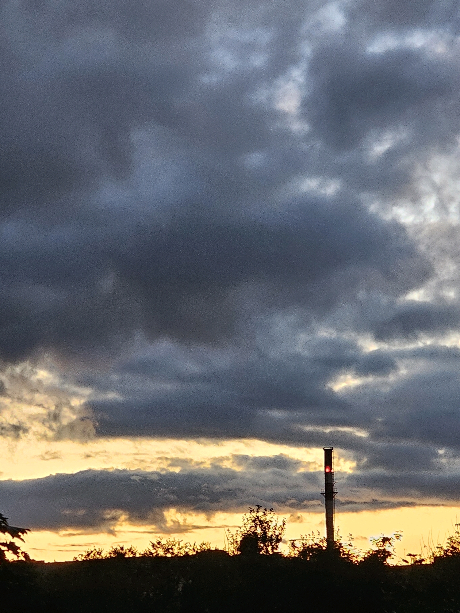 The image depicts a tall, industrial chimney with a red light illuminated near its top, standing against a dramatic sky filled with dark, heavy clouds. The horizon is a vibrant band of yellow and orange, suggesting either sunrise or sunset, with silhouetted trees and foliage in the foreground. The scene appears to be set in a somewhat industrial or rural area on the outskirts of Khimki, Russia, characterized by the prominent chimney and the natural, yet uncultivated, landscape. There are no people or animals visible in the frame, and no discernible actions or interactions are taking place. The overall atmosphere is one of subdued, possibly melancholic, beauty.