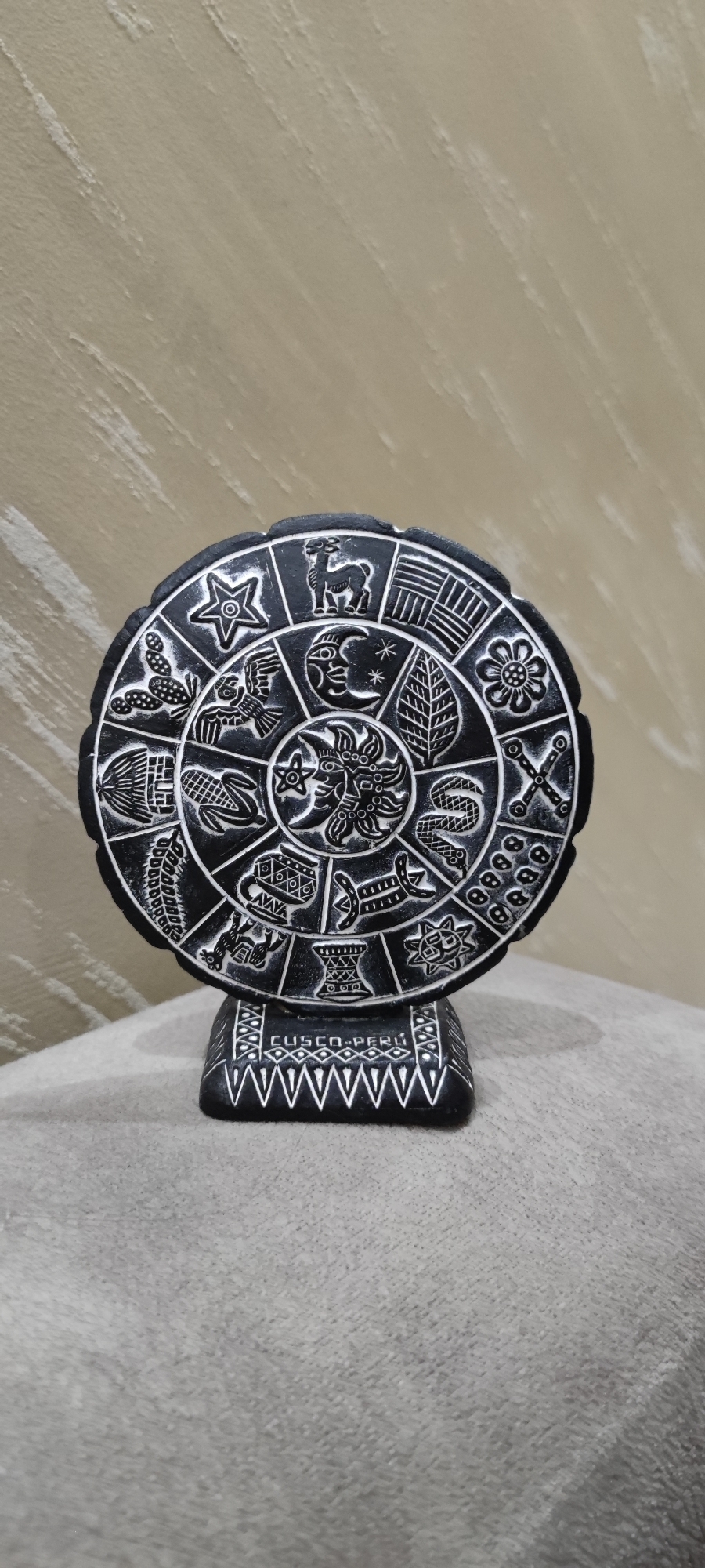 The image features a black, circular stone artifact resting on a gray, cushion-like surface. The artifact is engraved with a series of symbols and designs in a radial pattern, potentially representing a calendar or astrological chart. The artifact is supported by a rectangular base, on which the inscription "CUSCO - PERU" is visible. The background consists of a beige wall with subtle vertical striations. There is no indication that this image was taken in Yerevan, Armenia.