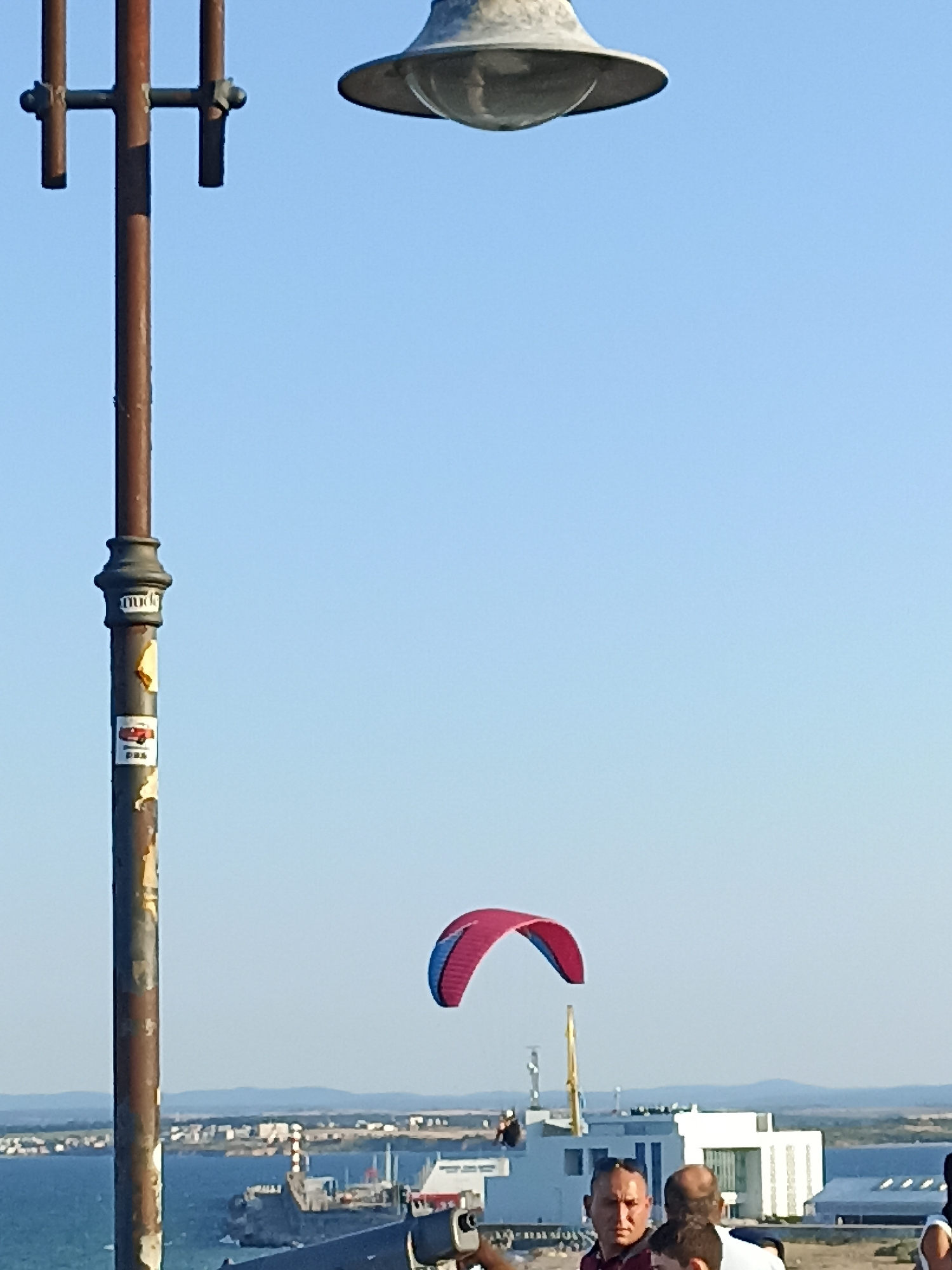 The image captures a sunny day in Burgas, Bulgaria, near the Sea Garden. In the foreground, a rusty lamppost with peeling paint stands tall, adorned with a few stickers. Above it, a retro-style lamp fixture is visible against a clear blue sky. Further in the background, a red and blue paraglider is suspended in the air, indicating recreational activity. Below the paraglider, a modern white building complex is situated near the sea, with various boats docked in the water. A group of people are gathered in the lower right portion of the image, appearing to observe the paraglider or engage in conversation. The overall scene suggests a leisurely day in a coastal urban environment.