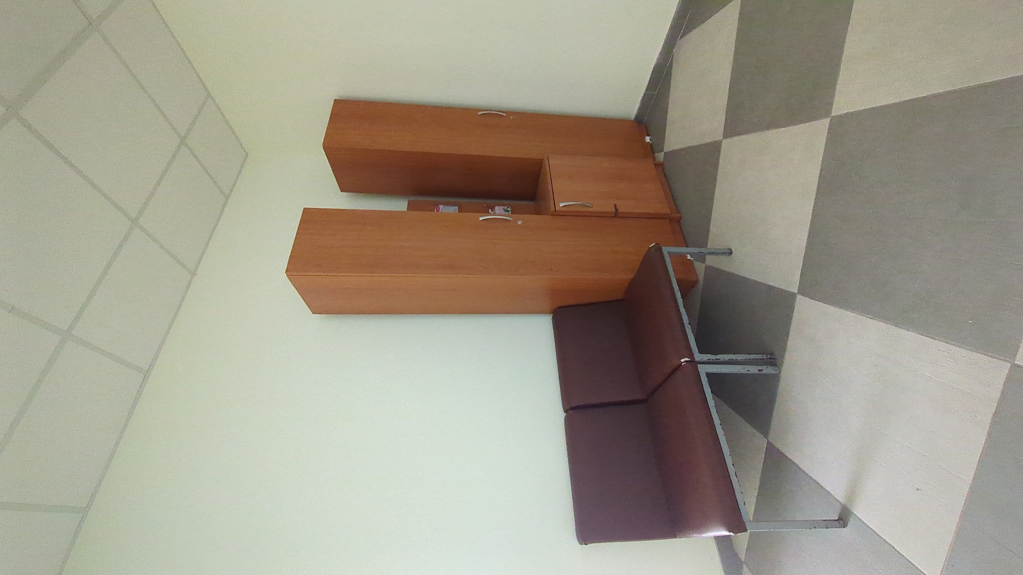 The image shows an empty waiting area with a two-seat bench and two tall wooden cabinets. The bench has dark brown upholstery and a metal frame. The cabinets are light brown and have silver handles. There is a smaller cabinet between the two tall cabinets. The floor is tiled with a checkered pattern of light gray and white squares. The walls are painted a light cream color, and the ceiling is a white drop ceiling with a grid pattern. The scene appears to be indoors, possibly in a public building or office. There are no people or any visible actions taking place.