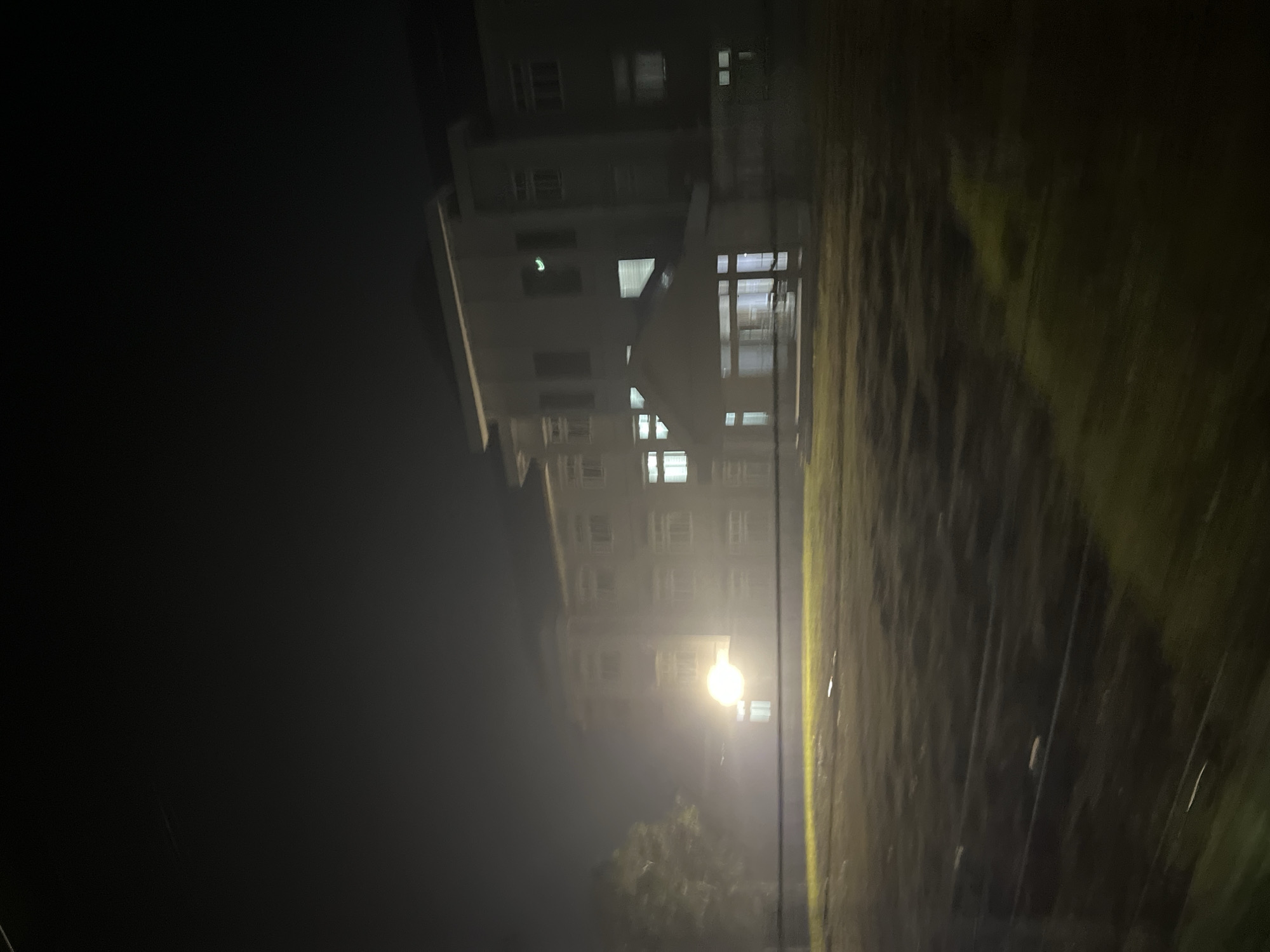 The image depicts a building at night, possibly a hospital, in the city of Kesehatanbandar, Indonesia. The building appears to be a multi-story structure with numerous windows, some of which are illuminated. A large, bright light source is visible to the left of the building, casting a glow on the surrounding area and a tree. The foreground shows a field or lawn, with some patches of illuminated ground. The scene is characterized by a dark sky and a slightly hazy atmosphere, contributing to a dimly lit and somewhat obscured view. The image appears to be taken from a moving vehicle, as evidenced by the motion blur.