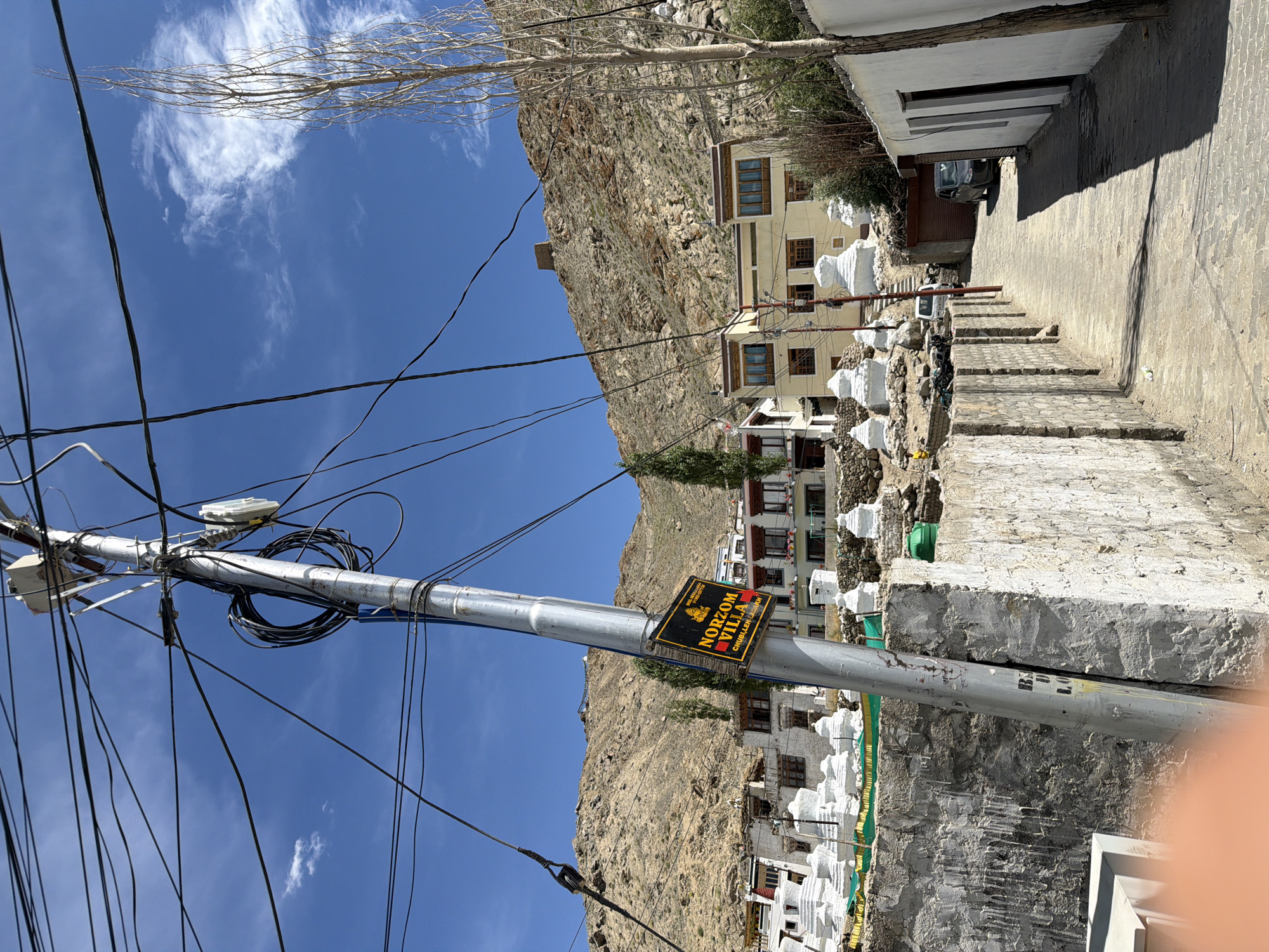 A street in Leh, India, features a dirt path leading past whitewashed buildings and stupas, set against a backdrop of arid mountains. A prominent utility pole with numerous tangled wires stands in the foreground, adorned with a sign indicating "Norzom Villa, Chushot, Leh." The scene is bathed in bright sunlight, casting shadows on the ground. No people are visible in the frame, but a parked car and a few motorcycles are present. The architecture suggests a traditional Ladakhi style.