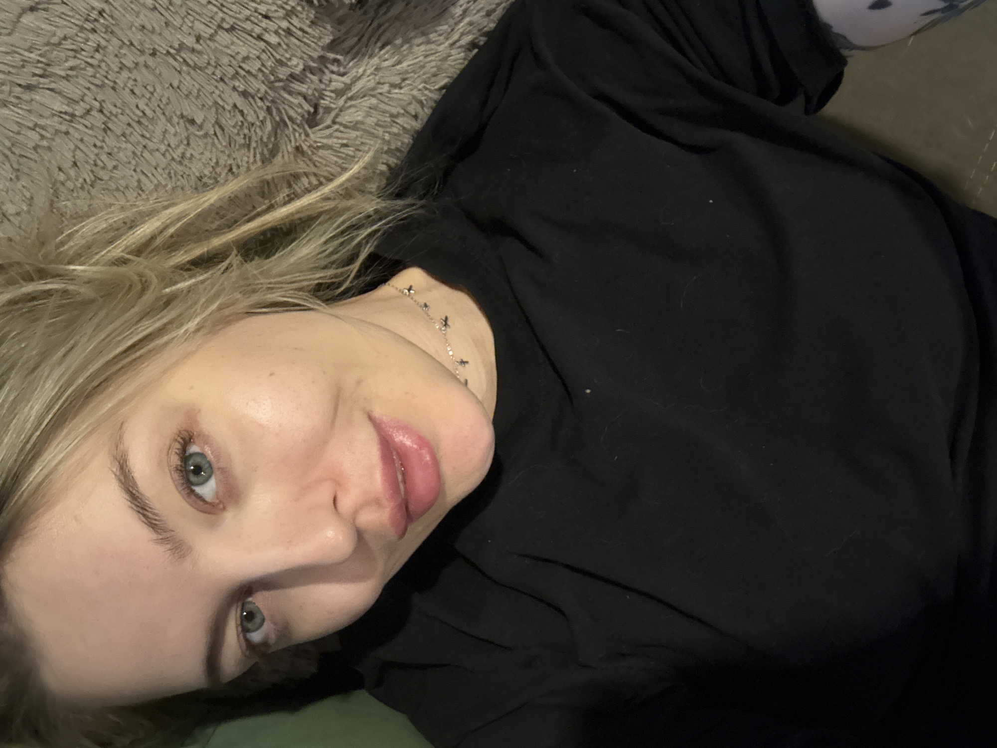 A pale-skinned woman with blonde hair and blue eyes is lying on a surface, possibly a bed or couch, covered with a gray, furry material. She is wearing a black long-sleeved shirt and a silver necklace with small pendants around her neck. The image is taken from a high angle, and the woman is looking directly at the camera with a slight smile. A tattoo is visible on her right arm. There is no indication of the city of Krasnoyarsk, Russia in the image.