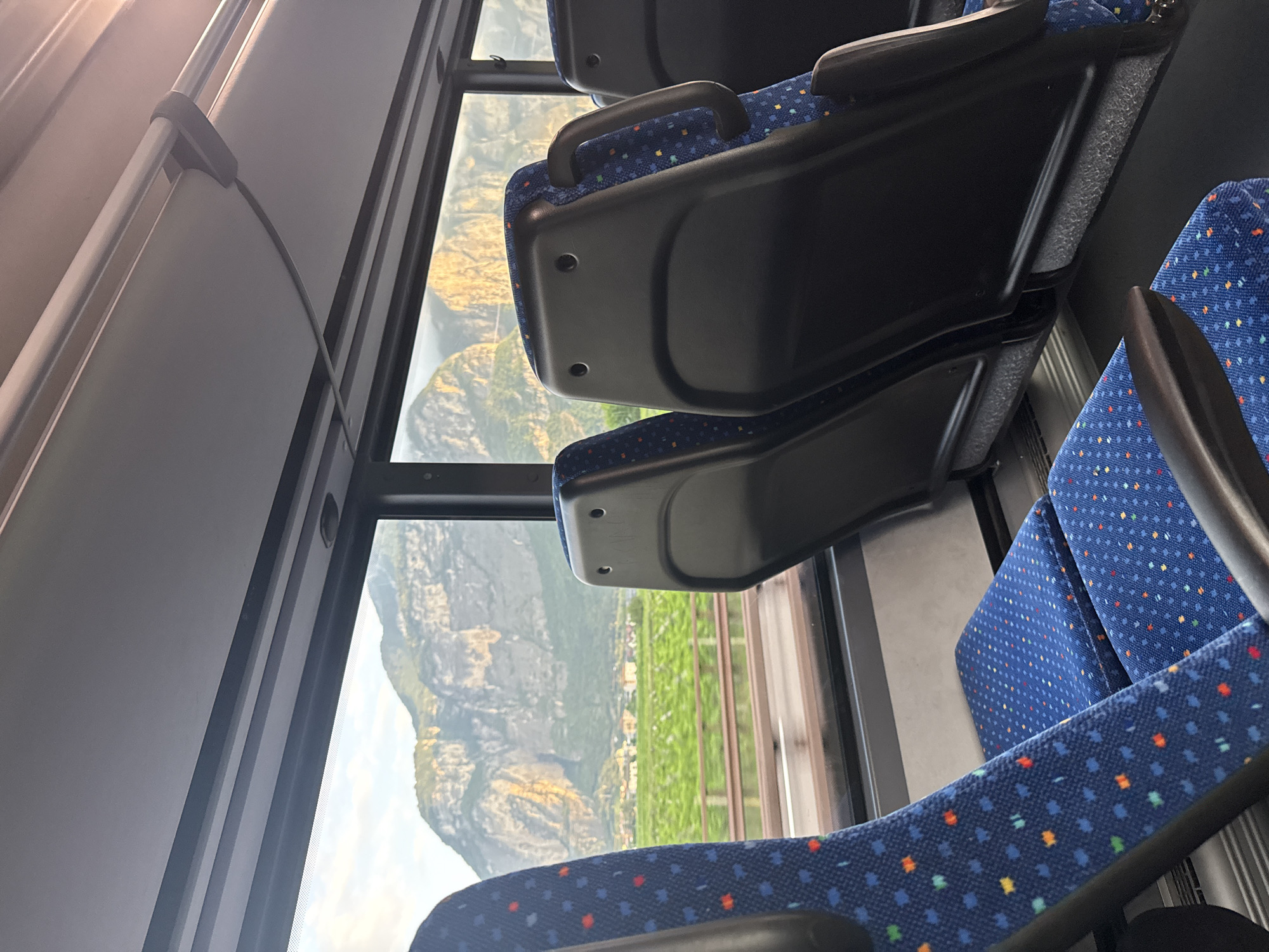 The image shows the interior of a bus with a view out of the window. In the foreground, two blue seats with colorful polka dots are visible. The seat on the left is partially in frame, while the seat on the right is more fully visible, showing its black headrest with a dark gray handle. Beyond the seats, a large window reveals a scenic landscape. Rugged, rocky mountains with patches of vegetation are bathed in golden sunlight. In the middle ground, a green field, possibly vineyards, stretches across the view, with a few buildings nestled at the base of the mountains. A railing or barrier runs horizontally in front of the field. The sky above the mountains is a pale blue with hints of clouds. The overall impression is that of a journey through a picturesque mountainous region. This scene is consistent with the landscape seen from a bus travelling through or near Mezzocorona, Italy, a town situated in the Adige River valley surrounded by mountains.