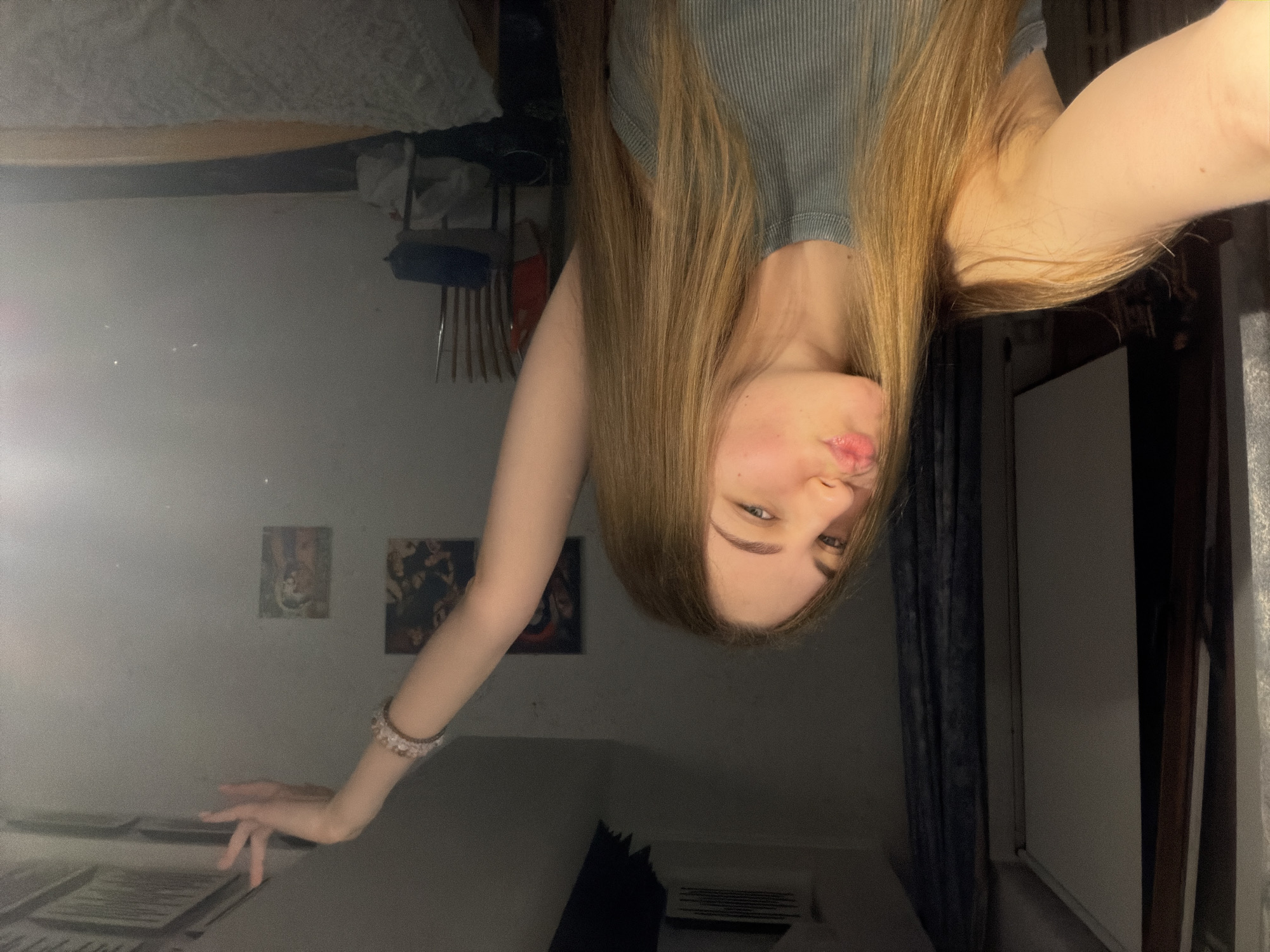 A young woman with long, blonde hair is taking a selfie in a dimly lit room. She is wearing a grey ribbed tank top and has a bracelet on her right arm, which is raised with her hand in a poised gesture. Her lips are pursed, and she is looking at the camera. In the background, there is a curtain, some framed pictures on the wall, and a bed with white bedding. The scene suggests a personal space, possibly a bedroom, in Dnipro, Ukraine.