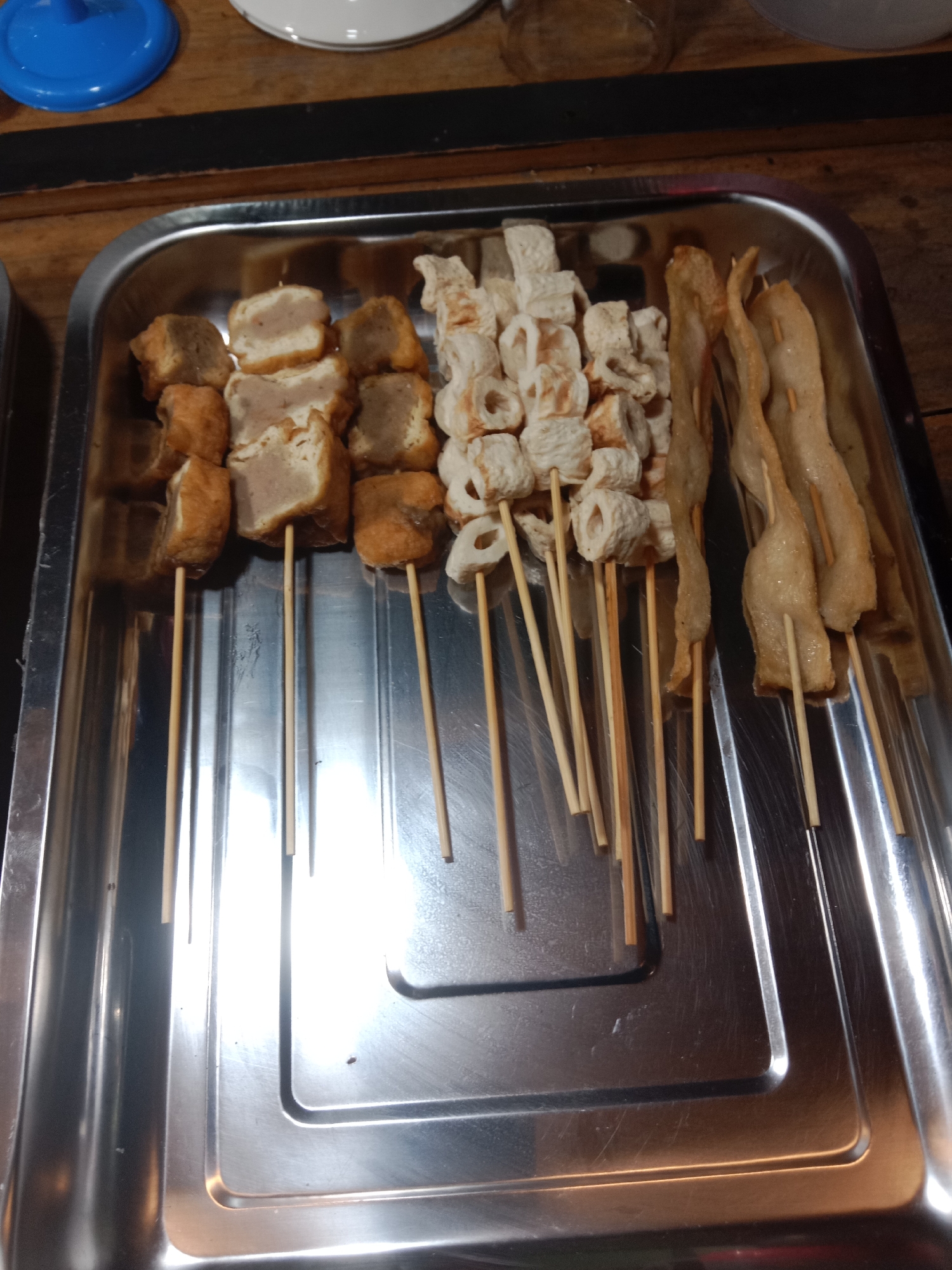 A stainless steel tray holds skewers of food. The tray is on a wooden surface. The food includes pieces of fried tofu and meat, and what appear to be fish cakes. The skewers are made of light-colored wood. The lighting suggests an indoor setting, possibly a food stall or market. The image appears to be taken at the Gatra Buana food stall, located in Lamongan, Indonesia.